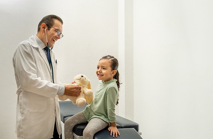A young girl sitting on an examination table, smiling at a doctor holding a teddy bear during a check-up in a medical office.