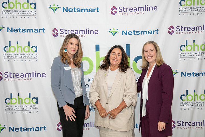 Three women stand together in front of a backdrop with logos for the County Behavioral Health Directors Association, Netsmart, and Streamline Healthcare. They are dressed in business casual attire and smiling.