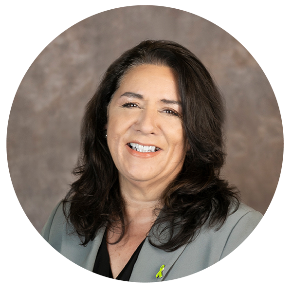 A smiling woman with dark brown hair, wearing a gray blazer with a yellow ribbon pin, against a textured gray background.