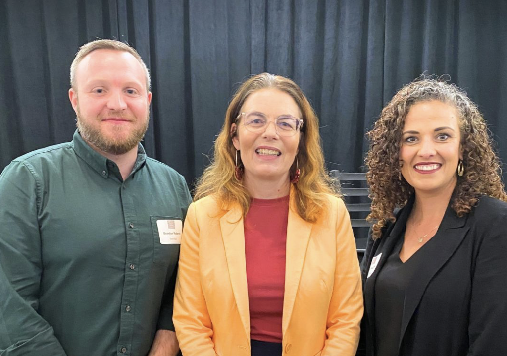 Three people smiling and standing close together in front of a black curtain, at a professional event or conference.