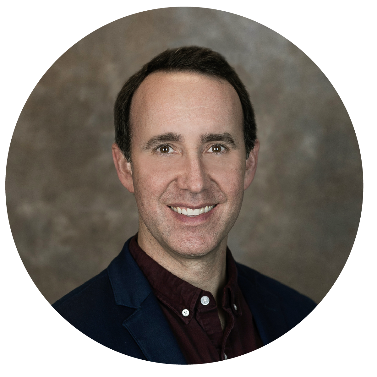 Headshot of a smiling man with short dark hair. He's wearing a dark blazer over a maroon shirt. The background is a blurred, textured brown.