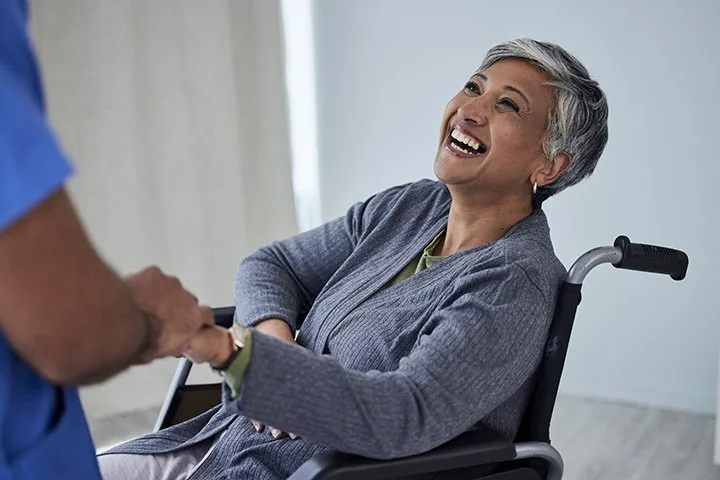 An elderly woman with gray hair smiling and shaking hands with a healthcare worker, sitting in a wheelchair in a bright room.