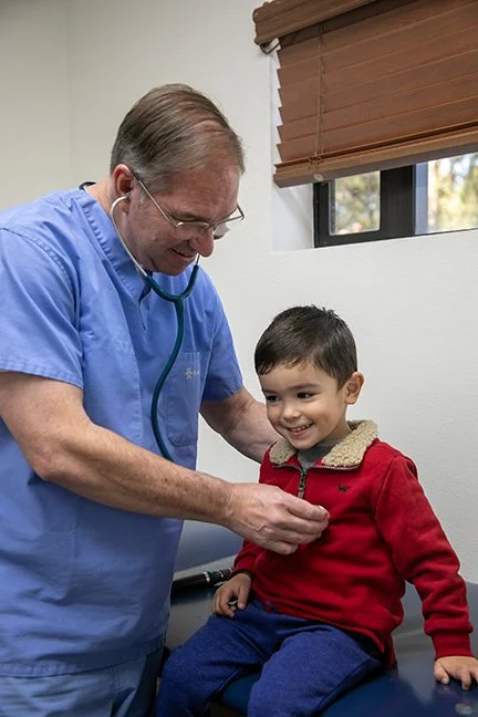 A male doctor in blue scrubs examining a young boy in a red jacket at a clinic.