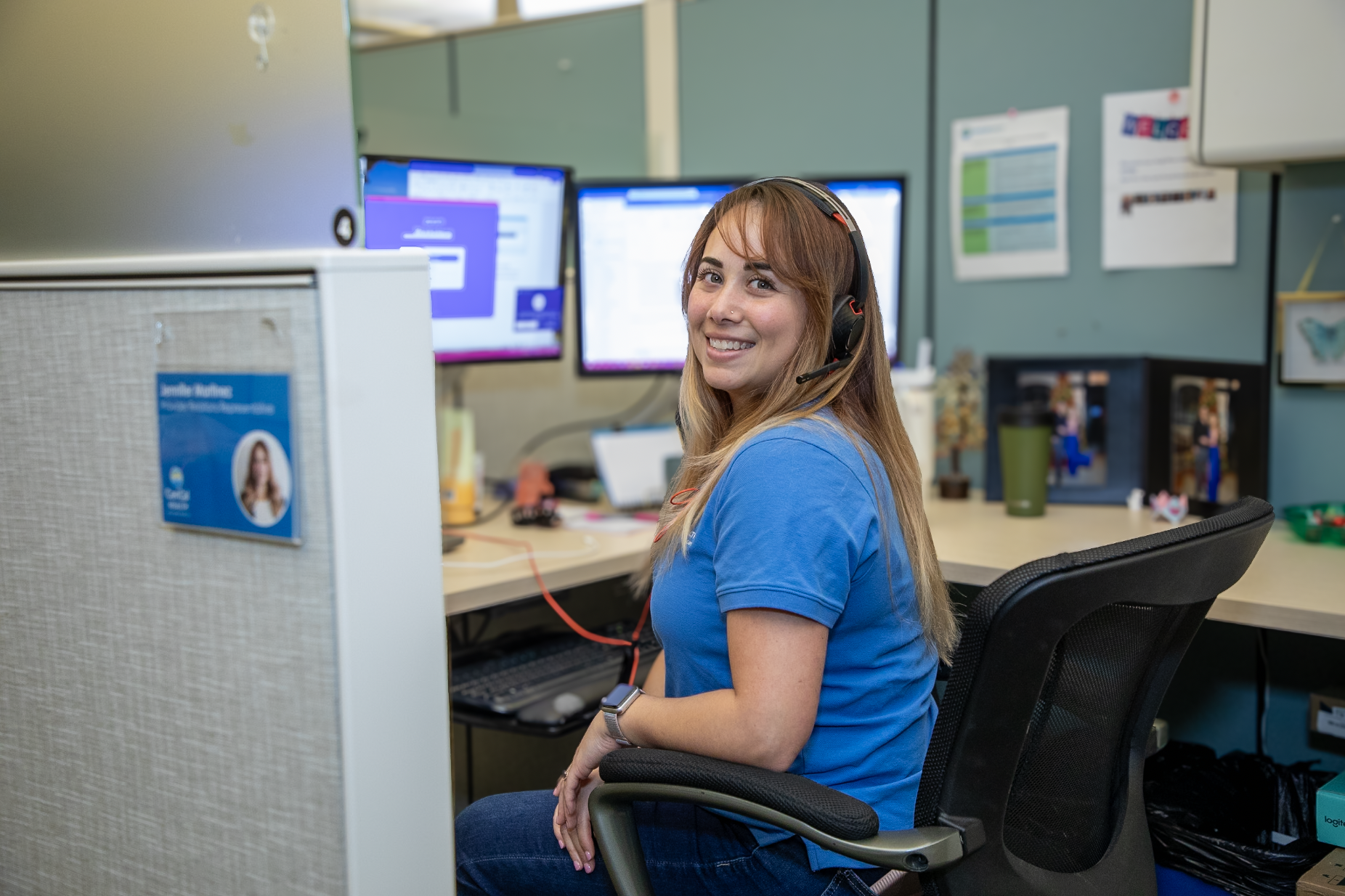 A smiling woman with long, light brown hair, wearing a blue shirt and a headset, sitting at a desk in an office with three computer monitors, a desktop computer, and various office supplies.