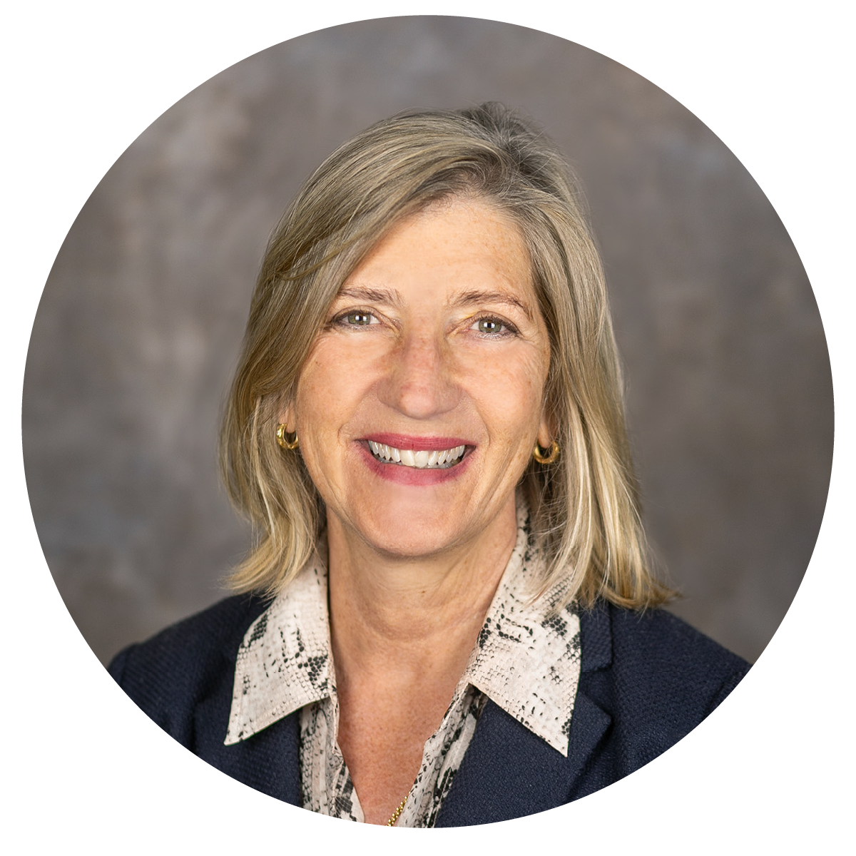 A smiling middle-aged woman with shoulder-length blonde hair, wearing a dark blazer and a patterned blouse, with gold hoop earrings, against a neutral background.