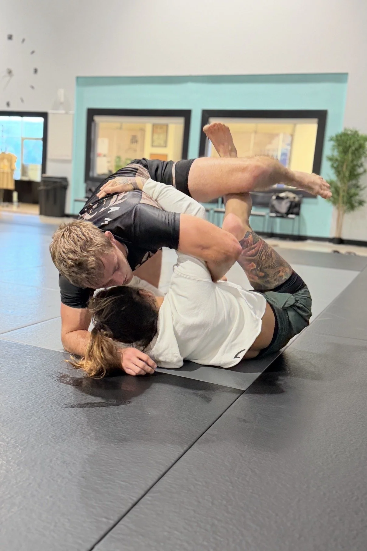 Two people practicing Brazilian Jiu-Jitsu on a mat, with one person on the bottom and the other on top, both engaged in a grappling position.