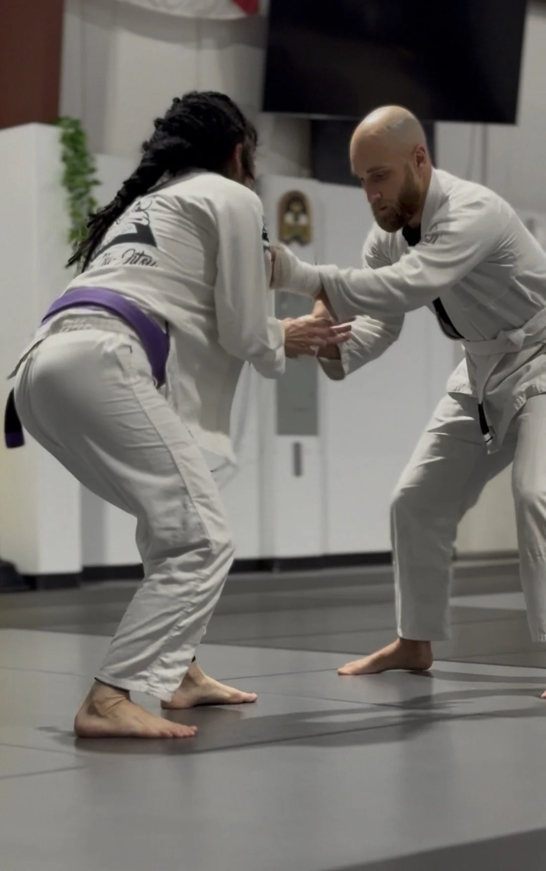 Two people practicing Brazilian Jiu-Jitsu in a gym, wearing gis, with one person with dreadlocks and the other bald, engaged in a grappling position.