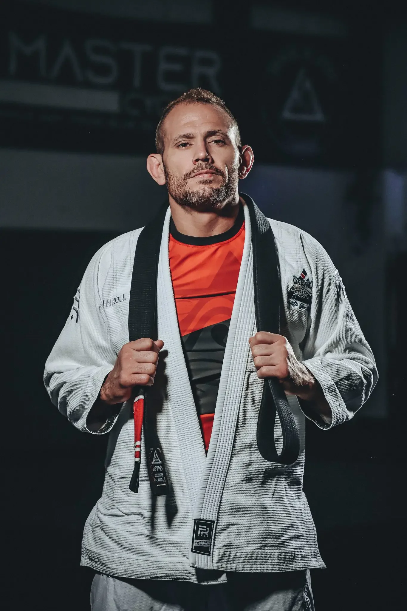 A confident male martial artist wearing a white gi with various patches, holding the lapels, standing in a gym, with a banner that says 'MARTER' in the background.