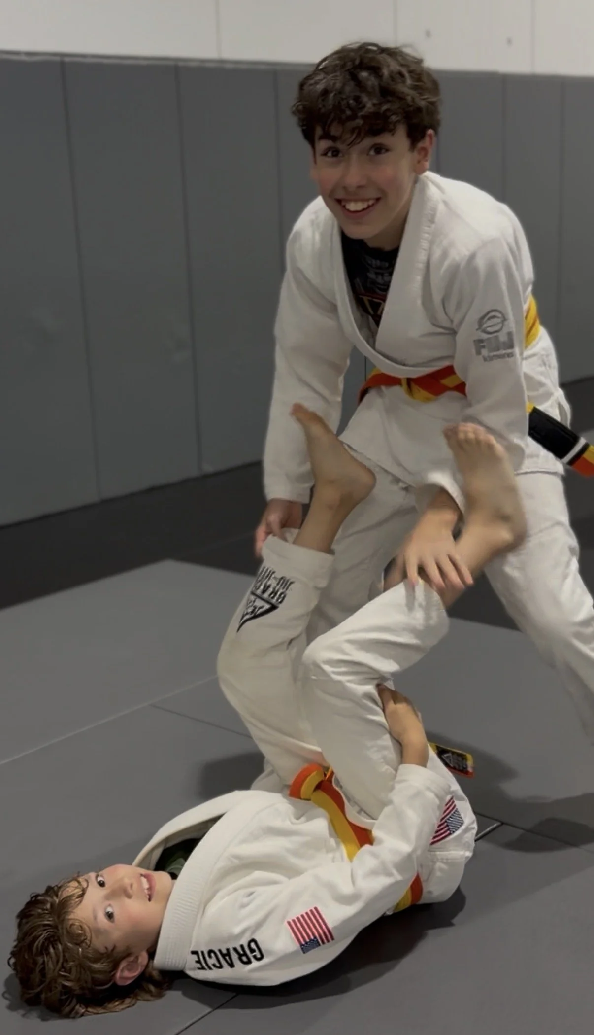 Kids practicing Brazilian Jiu-Jitsu in BJJ uniforms on a gray mat in a martial arts gym, engaging in a grappaling move, with smiling faces.
