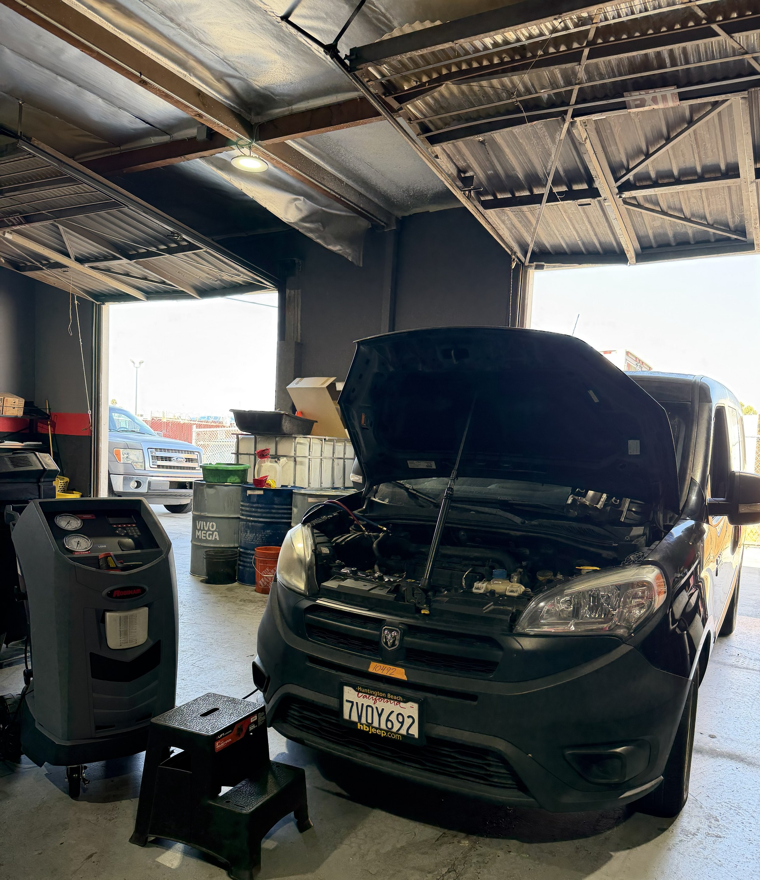 Auto repair shop with a black truck having its hood open, mechanical tools, and equipment around, including a diagnostic machine and car fluids, with an open garage door and other vehicles outside.