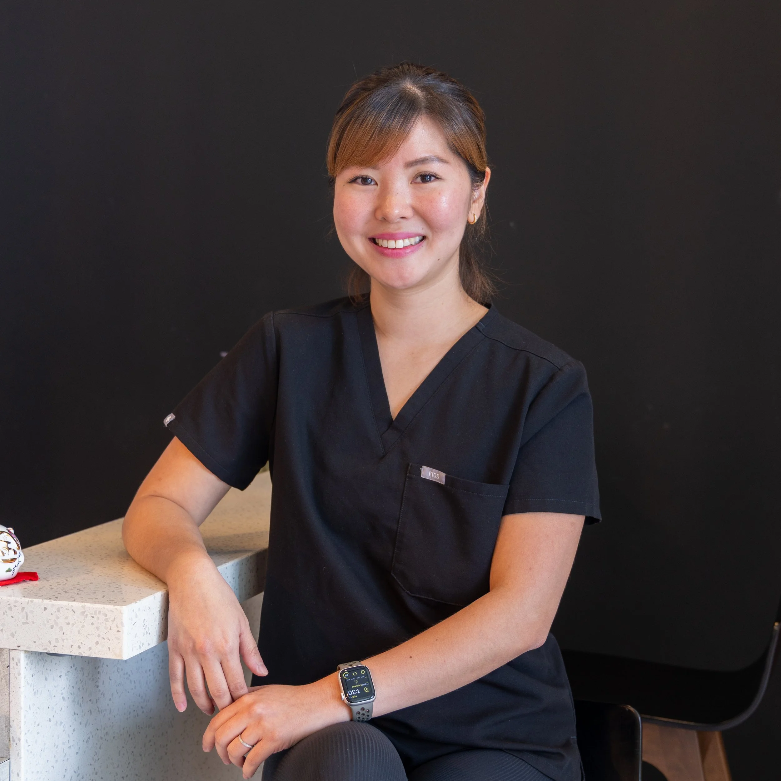 A woman in black scrubs sitting at a white speckled counter, smiling at the camera, wearing a smartwatch and a wedding ring, with a plain dark background.