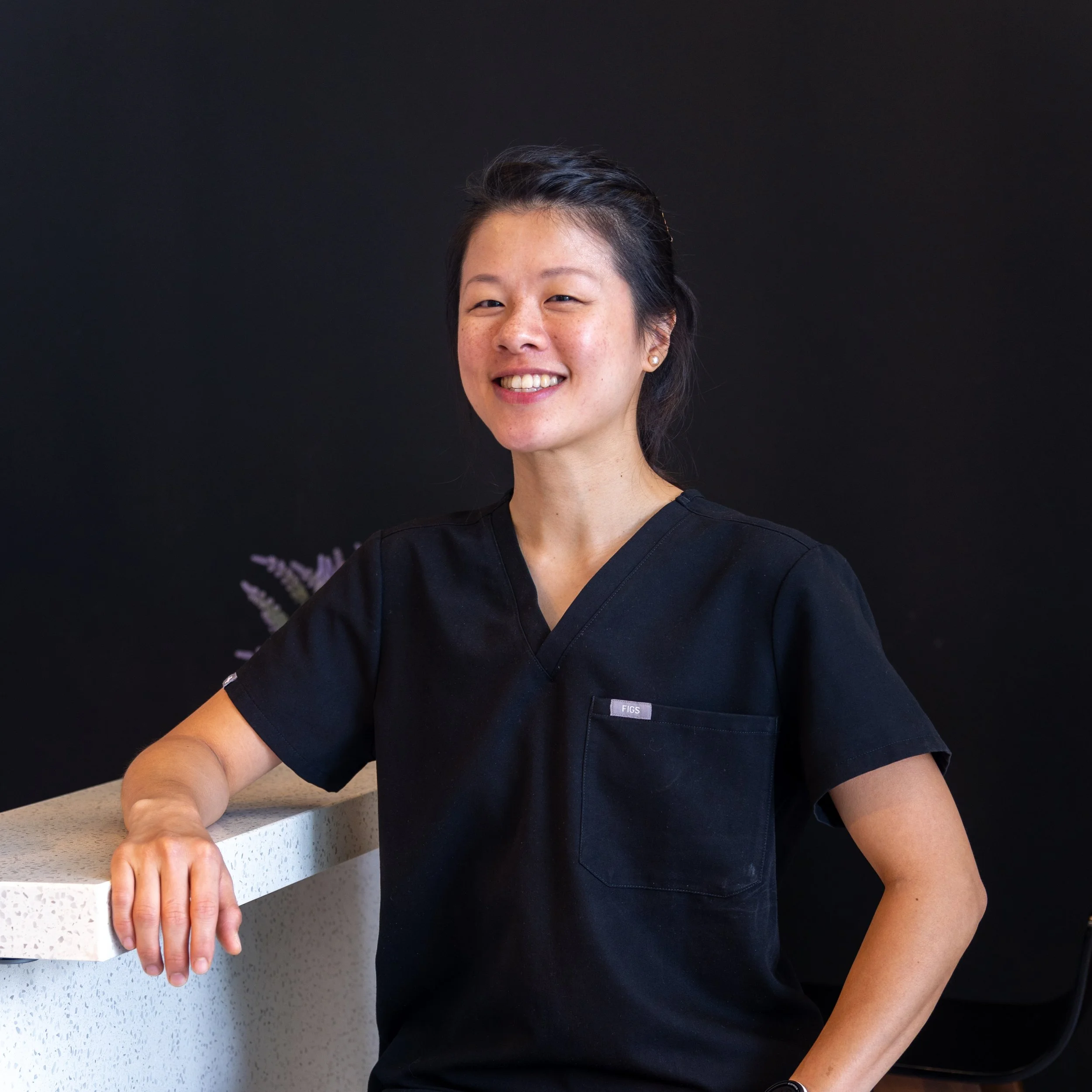 A woman in a black uniform smiling and leaning on a white speckled countertop against a black background.