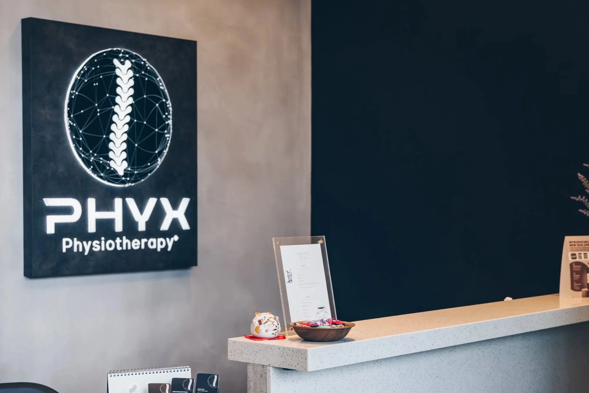 Reception area of PHYX Physiotherapy clinic with a illuminated sign featuring a spine graphic, a white countertop with a bowl of candies, a small decorative item, and a framed document.