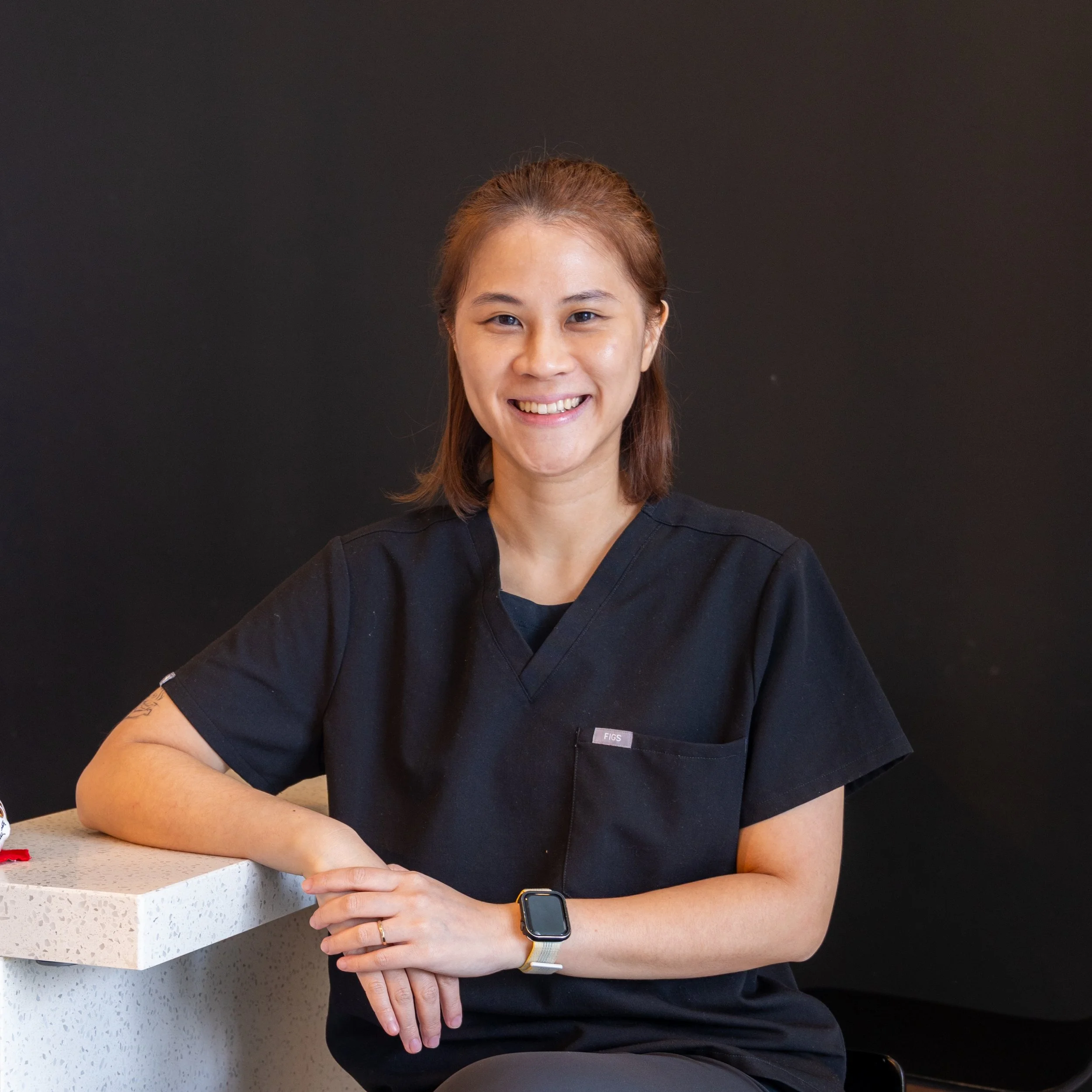A smiling woman with short brown hair wearing black scrubs and a smartwatch, sitting and resting her arm on a white speckled countertop against a black background.