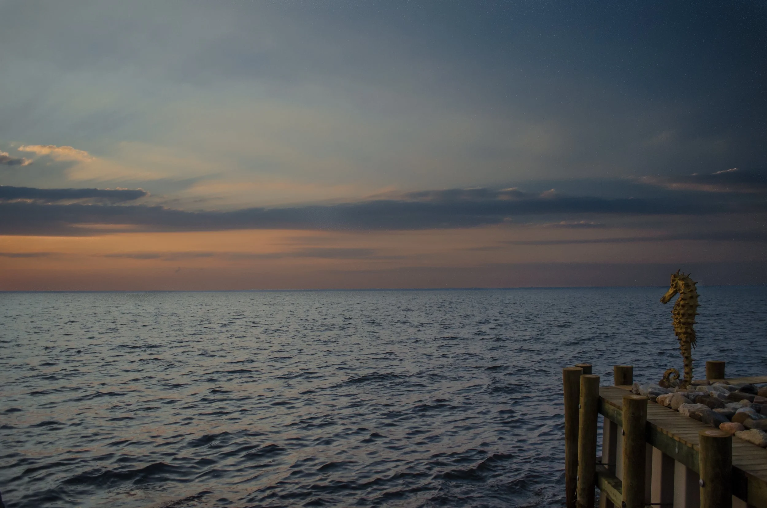 A seahorse sculpture on a rocky shoreline near a dock, with the ocean and a colorful sunset sky with clouds in the background.