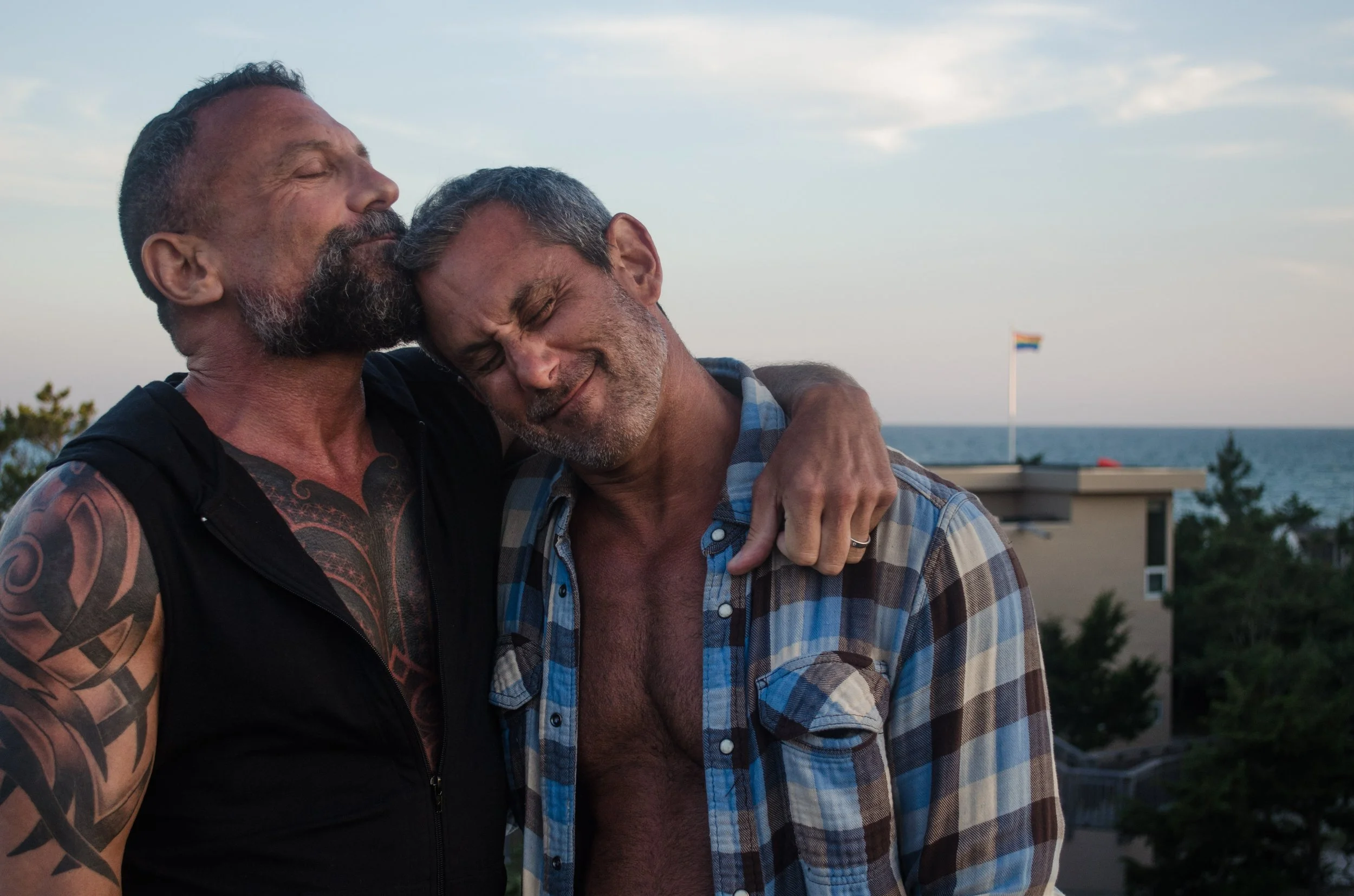 Two middle-aged men are embracing each other with closed eyes and content expressions; one has a beard, tattoos, and is wearing a black sleeveless vest, the other has gray hair and a beard, wearing a partially unbuttoned plaid shirt. The background s