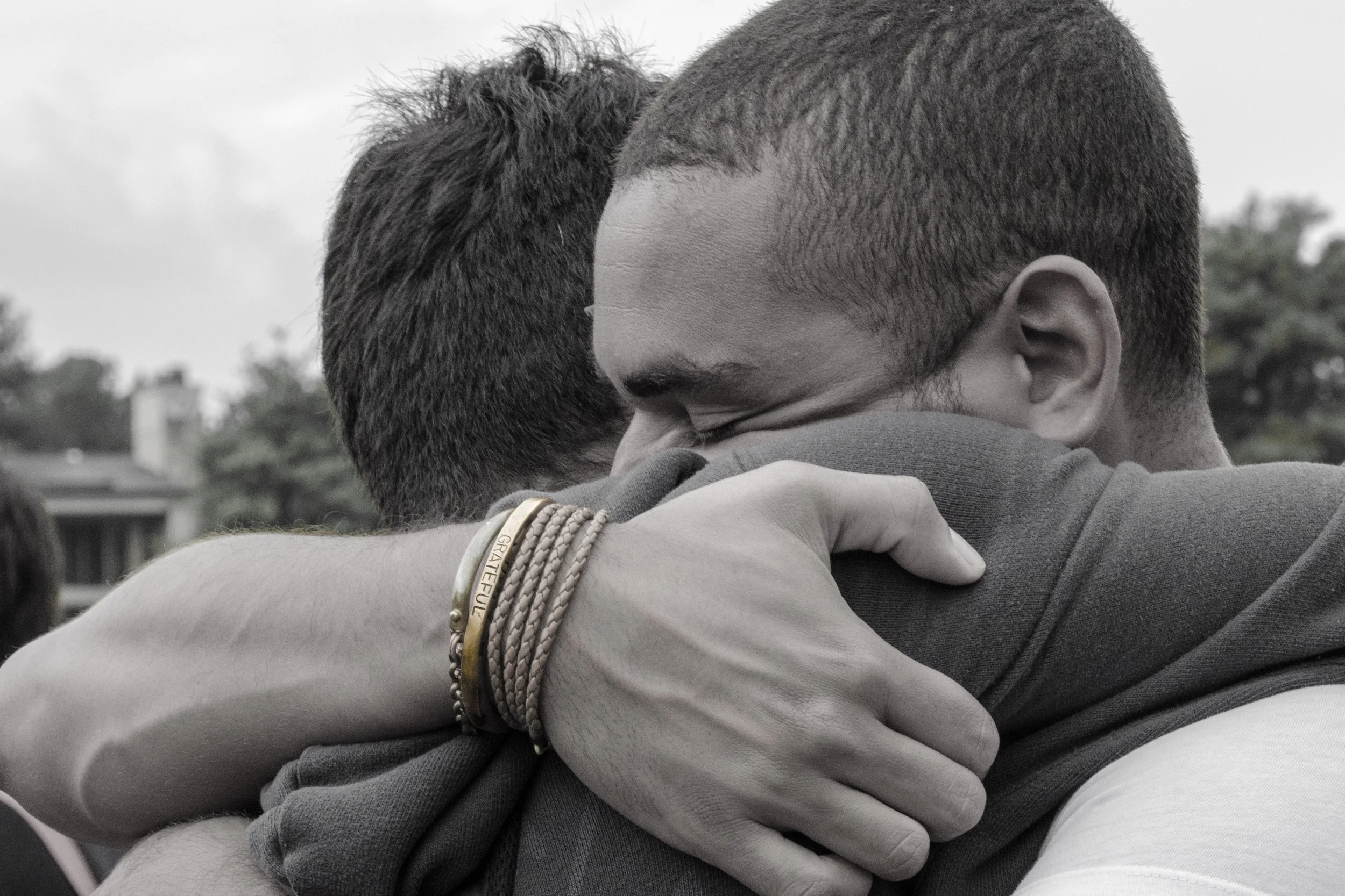 Two people hugging each other with their faces close together, one with short hair and the other with dark hair, one wearing multiple bracelets.