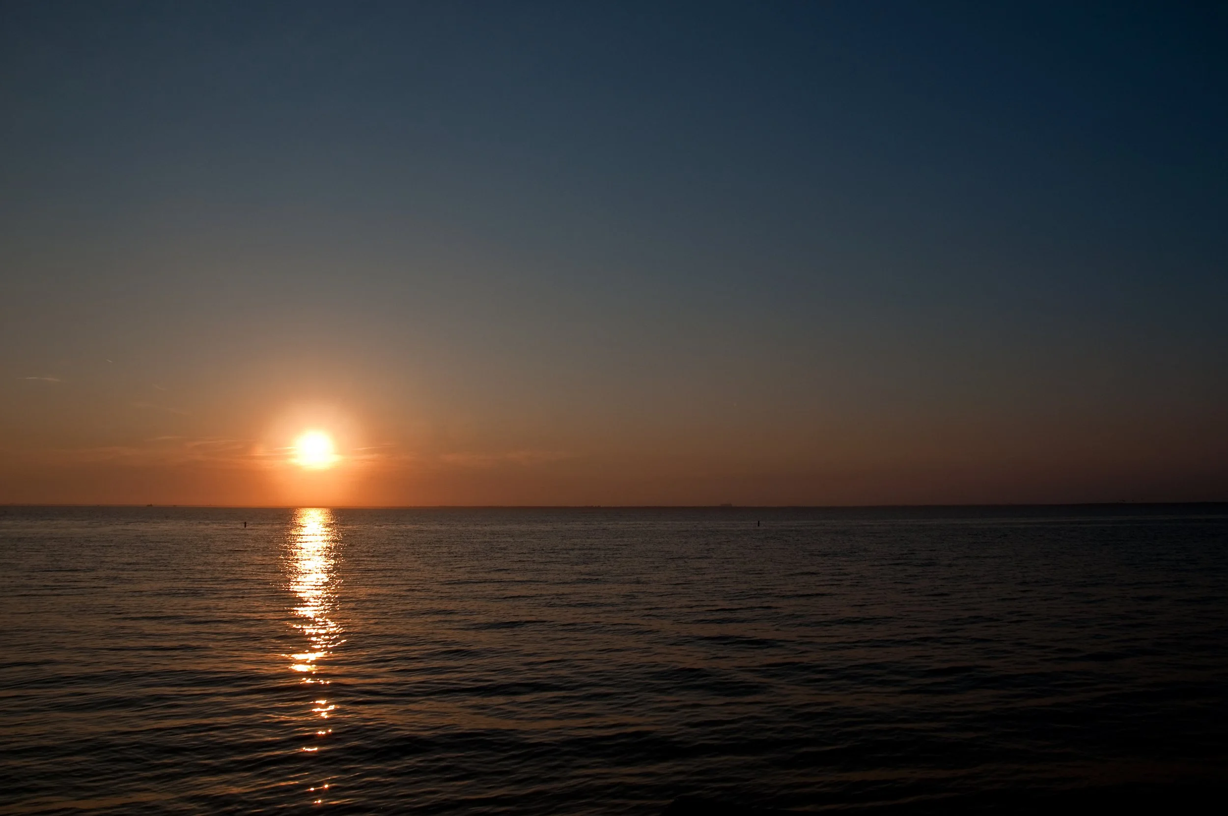Sunset over the ocean with reflected sunlight on the water, calm waves, and a clear sky.