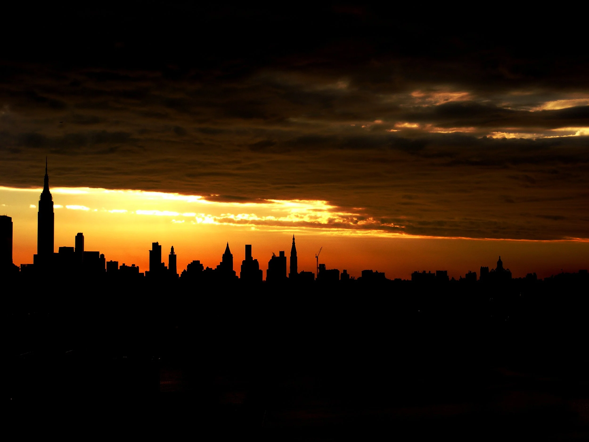 Silhouette of New York City skyline at sunset with dark clouds overhead and orange glow in the sky.