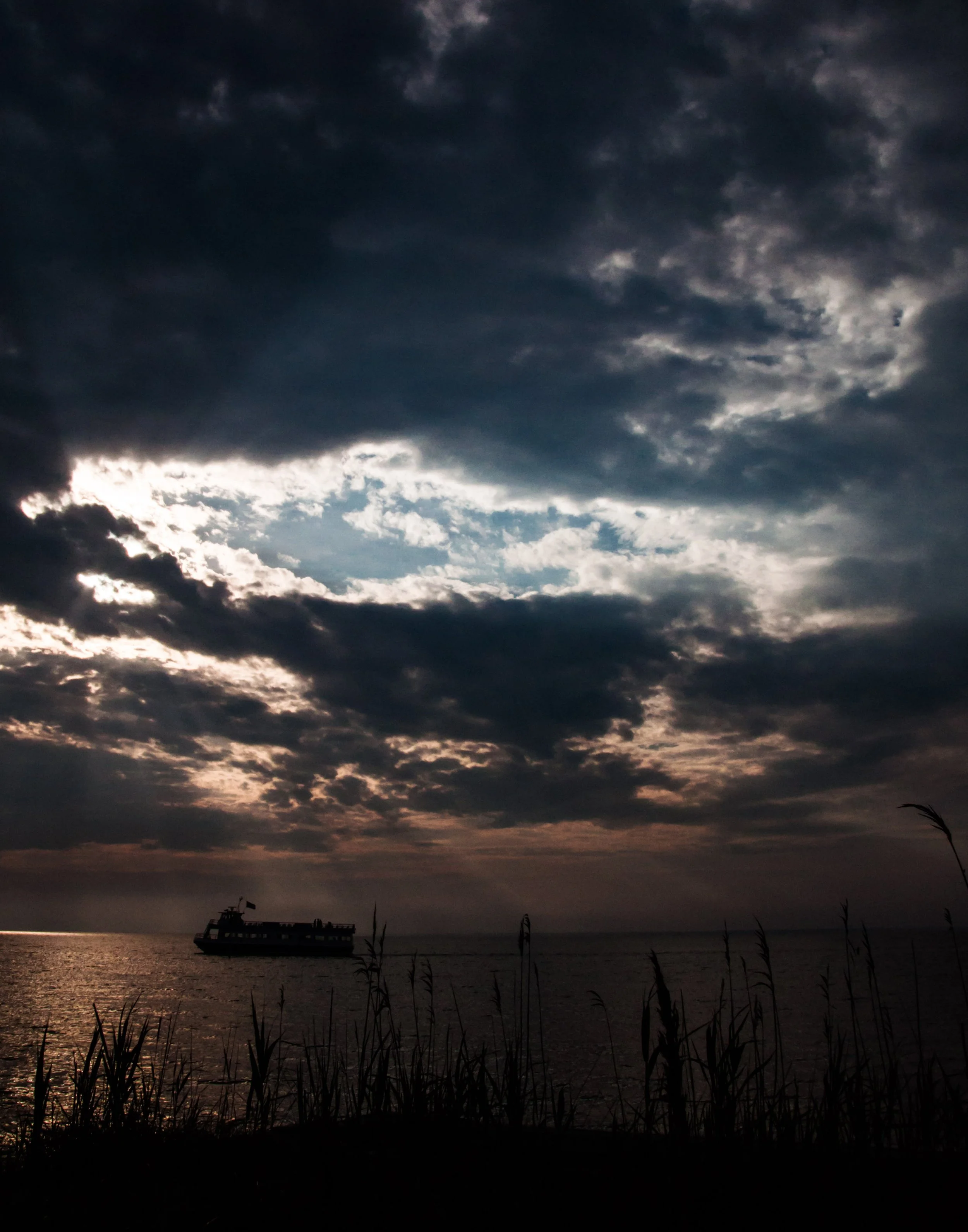 Dark cloudy sky over a body of water with a boat in the distance, silhouetted grass in the foreground.