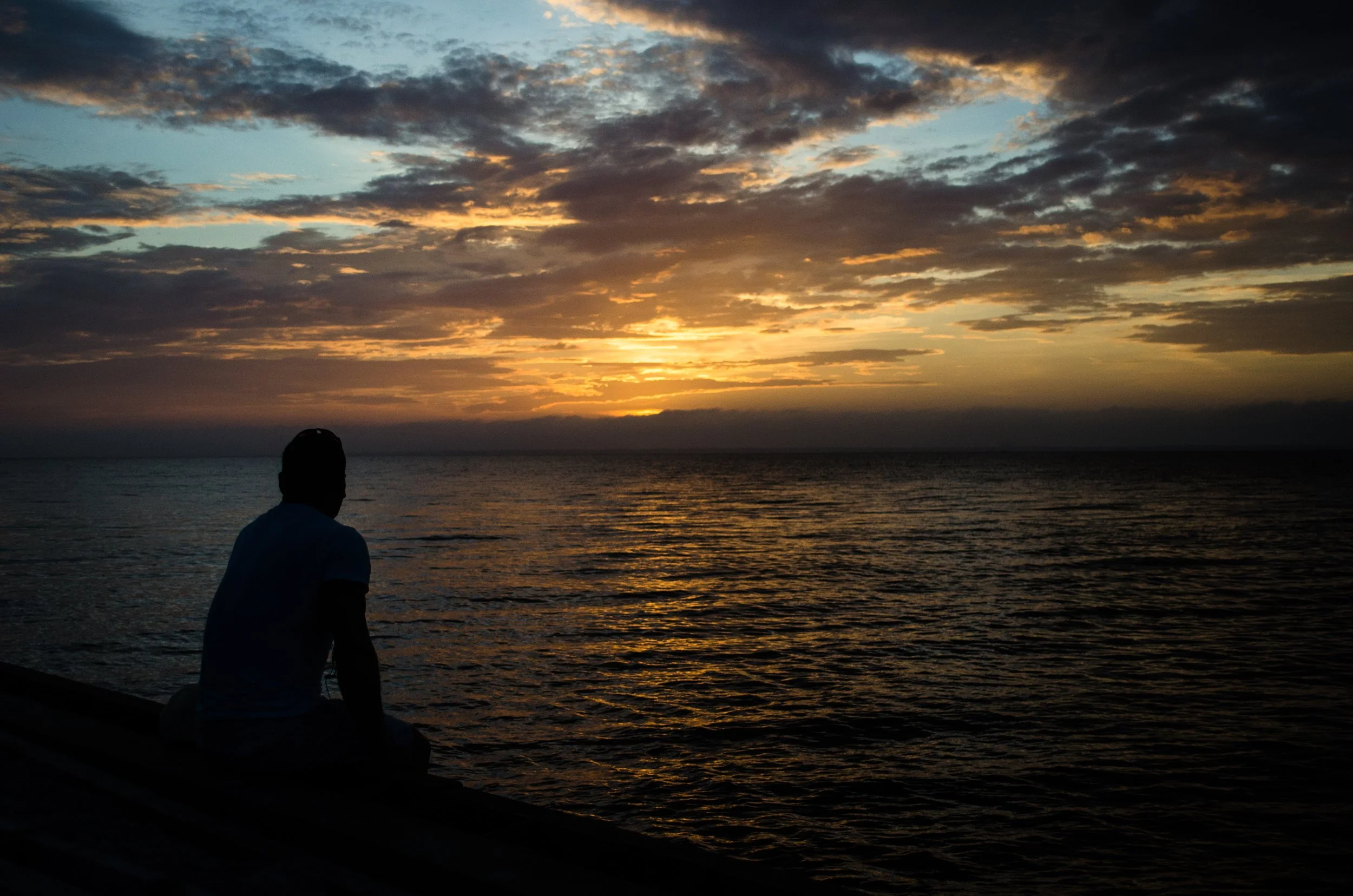 Silhouette of a person sitting by the water at sunset, with the sky filled with clouds and the horizon glowing with the setting sun.