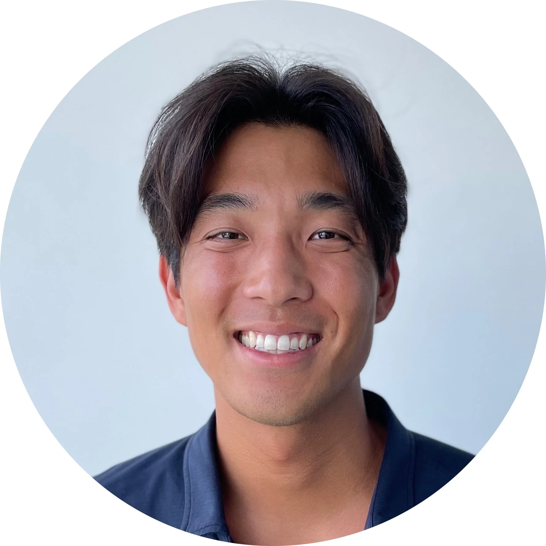 A close-up portrait of a smiling young man with dark hair, wearing a dark blue shirt, against a plain, light-colored background.