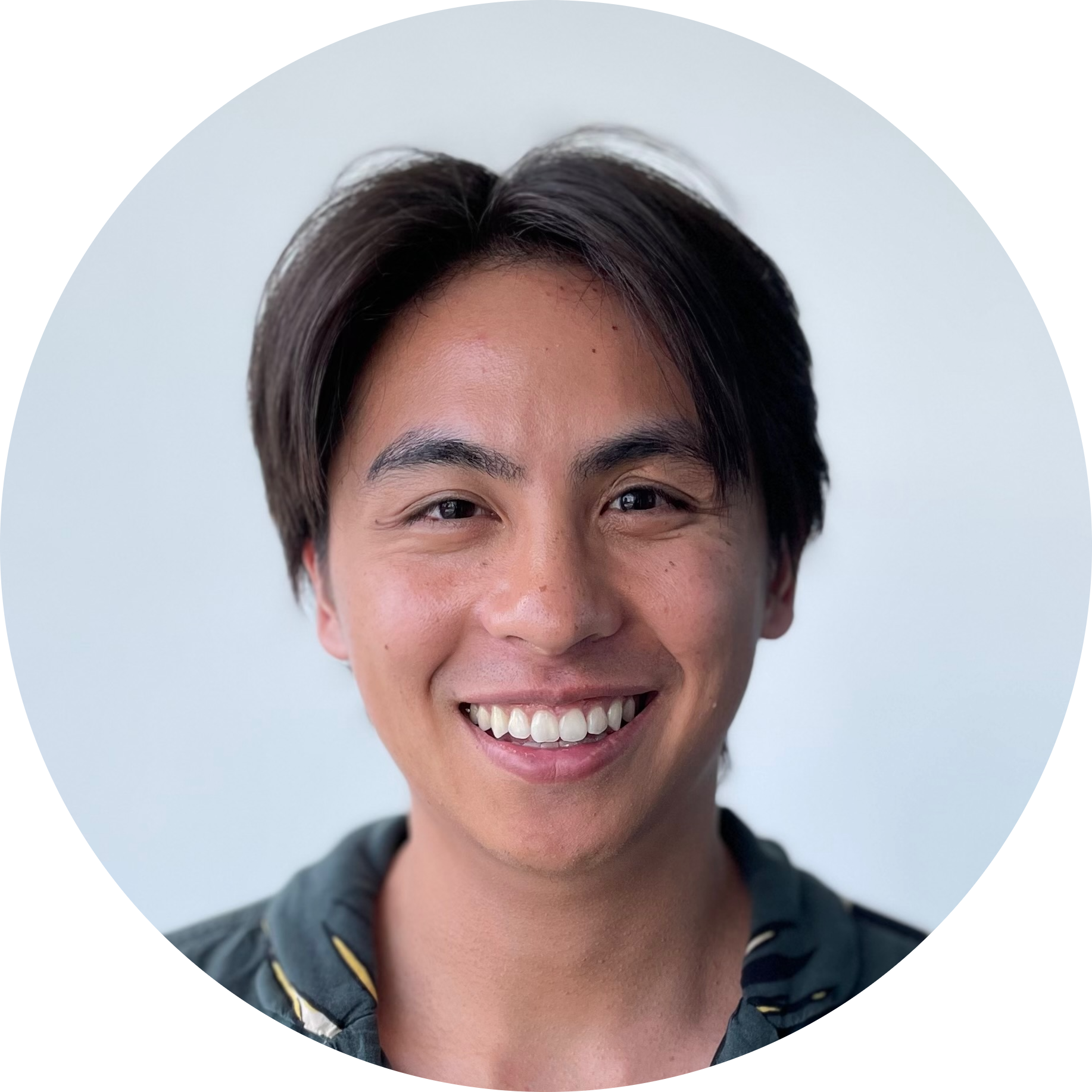 A young man with dark hair and a bright smile wearing a dark hoodie over a patterned shirt, standing against a light-colored background.