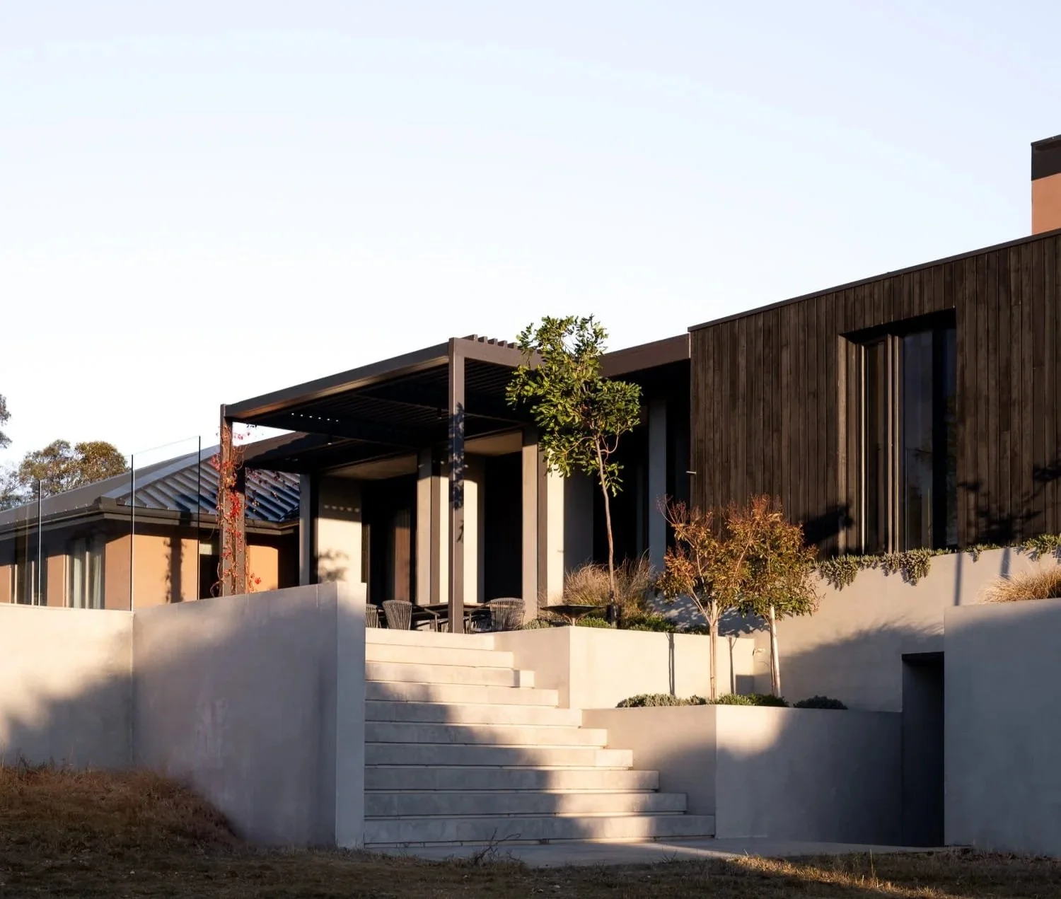 Modern house with dark wooden exterior, large windows, and a patio with chairs and tables, surrounded by retaining walls and landscaping, under clear sky.