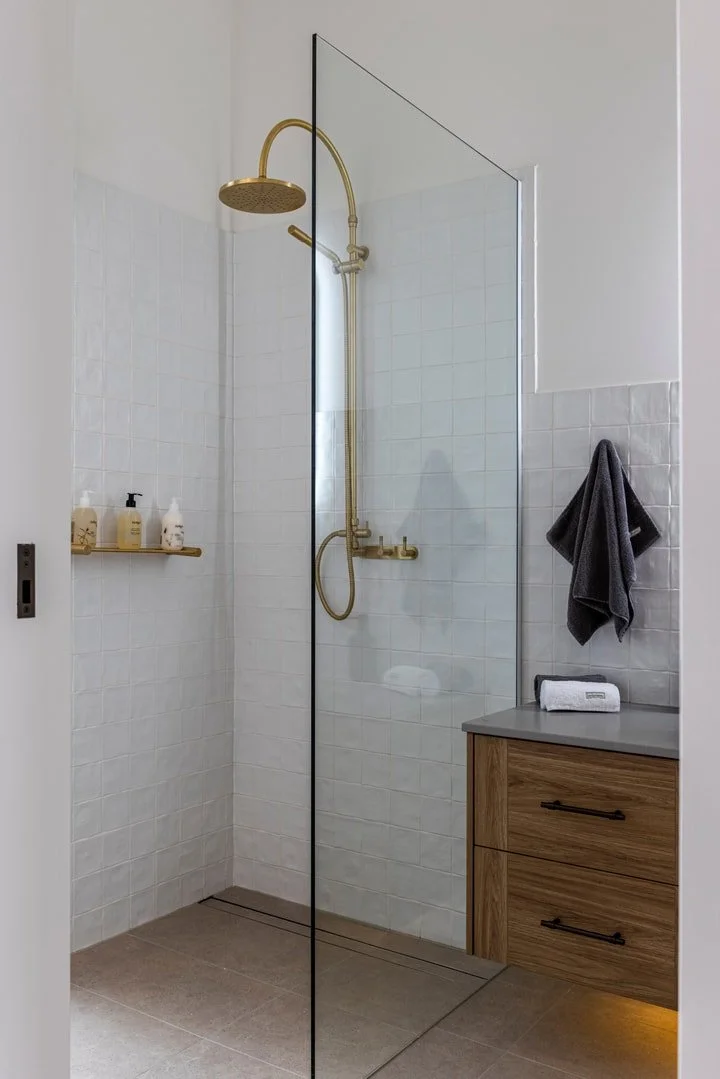 Modern bathroom with walk-in shower featuring gold fixtures and a glass partition, grey countertop with a wooden cabinet, dark towel hanging on wall, and a small shelf with toiletries.