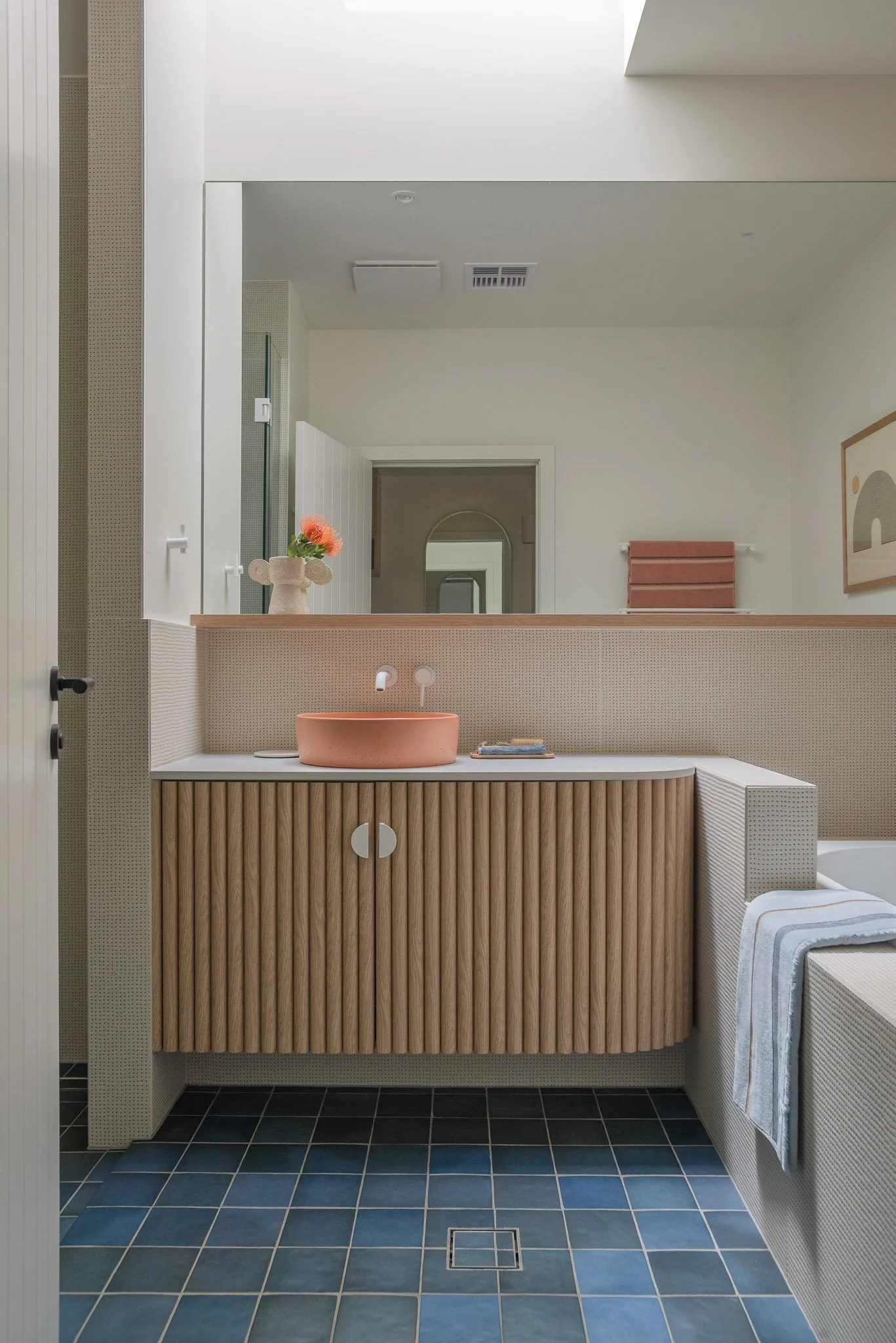 Modern bathroom with a pink vessel sink, a vase with pink flowers, a mirror, blue tiled floor, and a striped towel on a white textured bench.