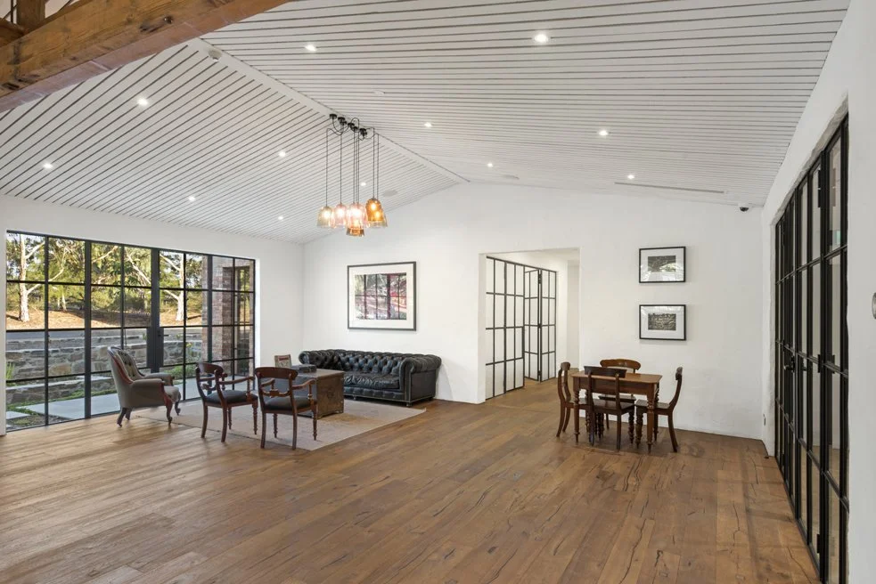 Spacious modern living room with large glass doors, wooden flooring, white walls, a black leather couch, and various chairs and art on the walls.