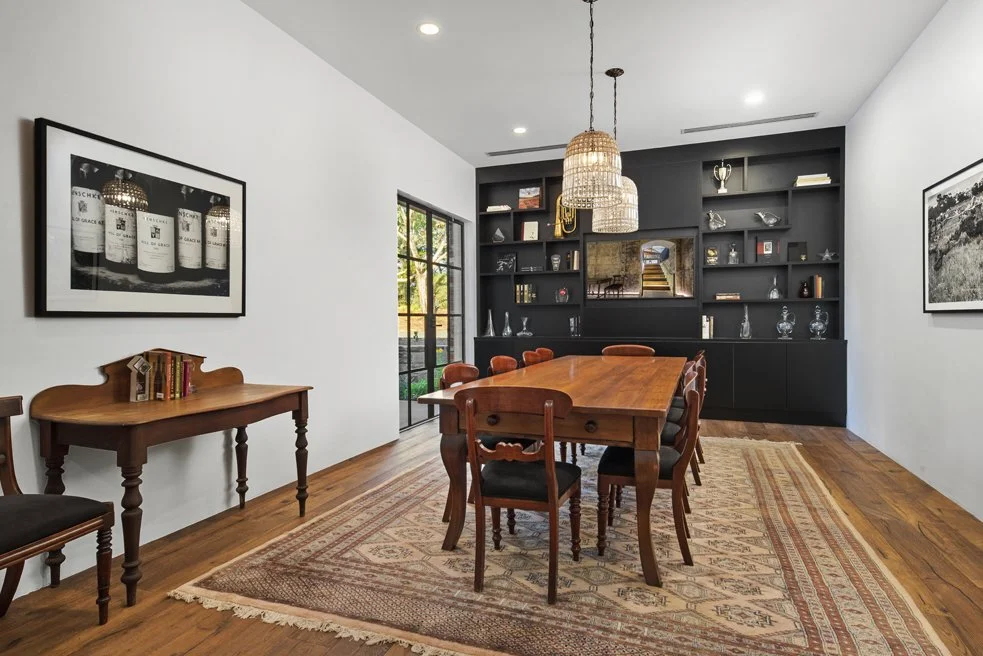 Dining room with a wooden table and chairs, black built-in shelves with decor, a large window, and framed art on white walls.