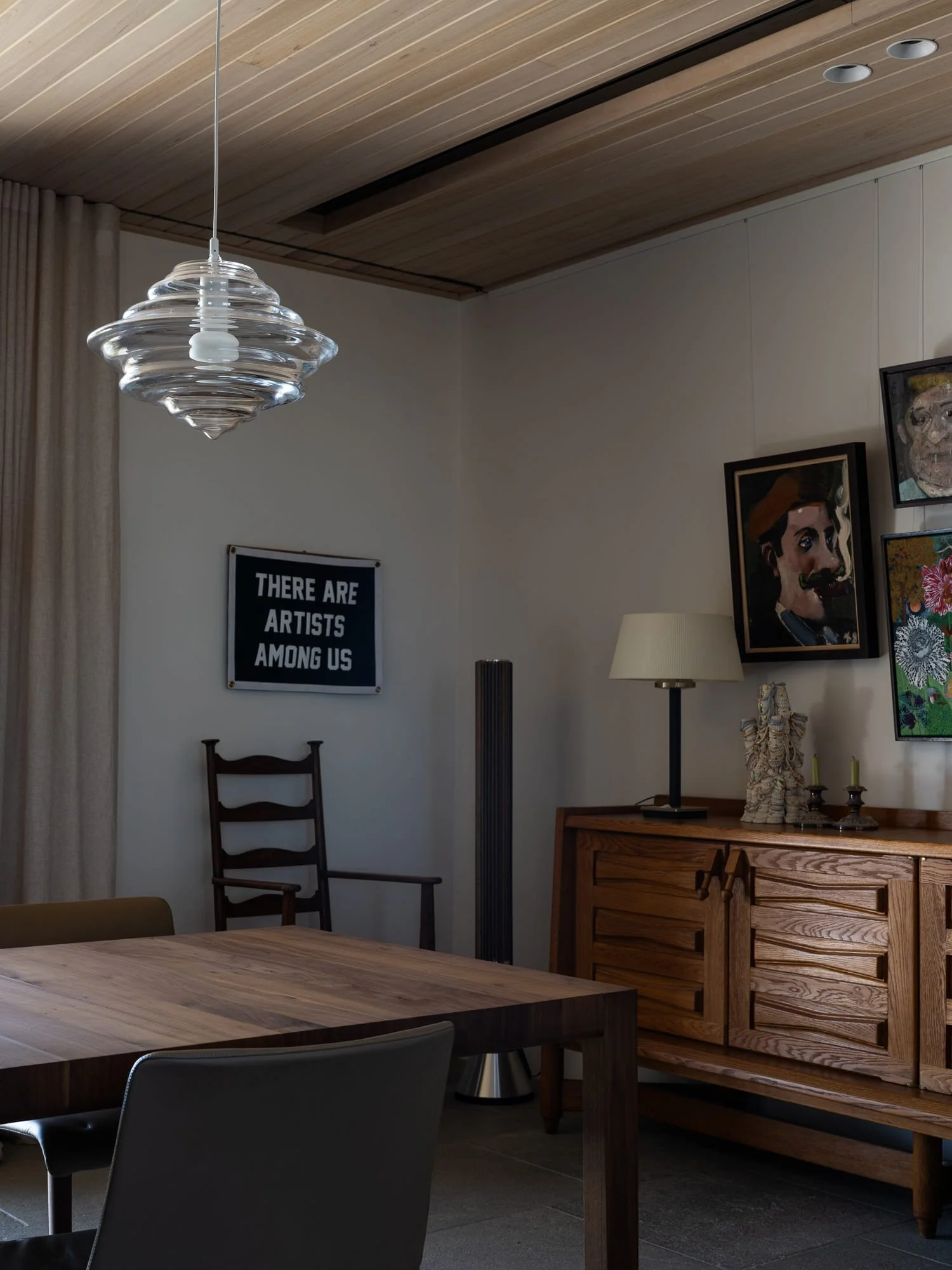 Interior of a room with wooden ceiling, artwork on the walls, a wooden sideboard, a table, and chairs, with a pendant light hanging from the ceiling.