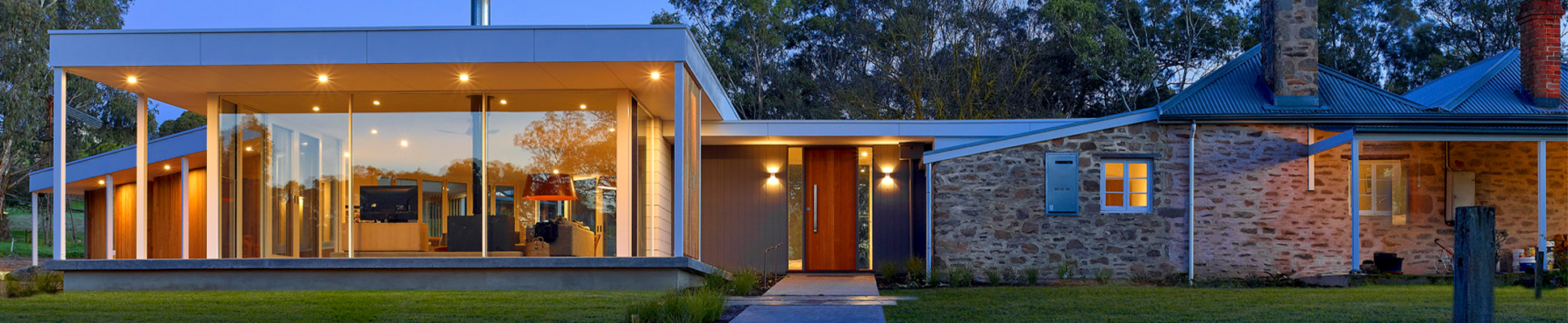 Modern house with large glass windows, stone and wood exterior, and a spacious lawn, illuminated during twilight.