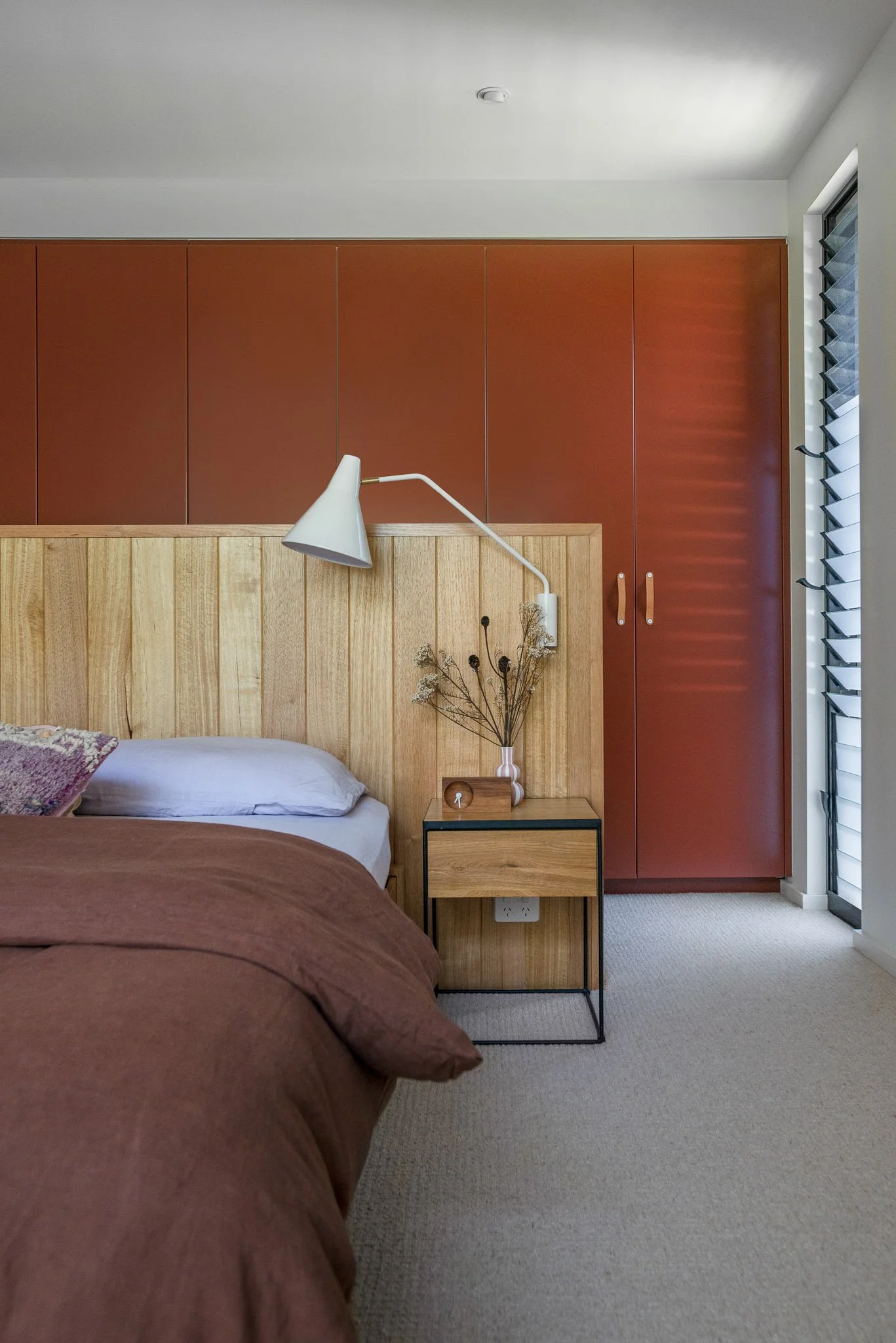 A bedroom with a bed that has maroon bedding, a wooden headboard, and a pillow. Next to the bed is a bedside table with a small clock, a vase with dried flowers, and a white wall-mounted lamp. There are wooden cabinets above the headboard and a window with blind louvered shutters on the right side.