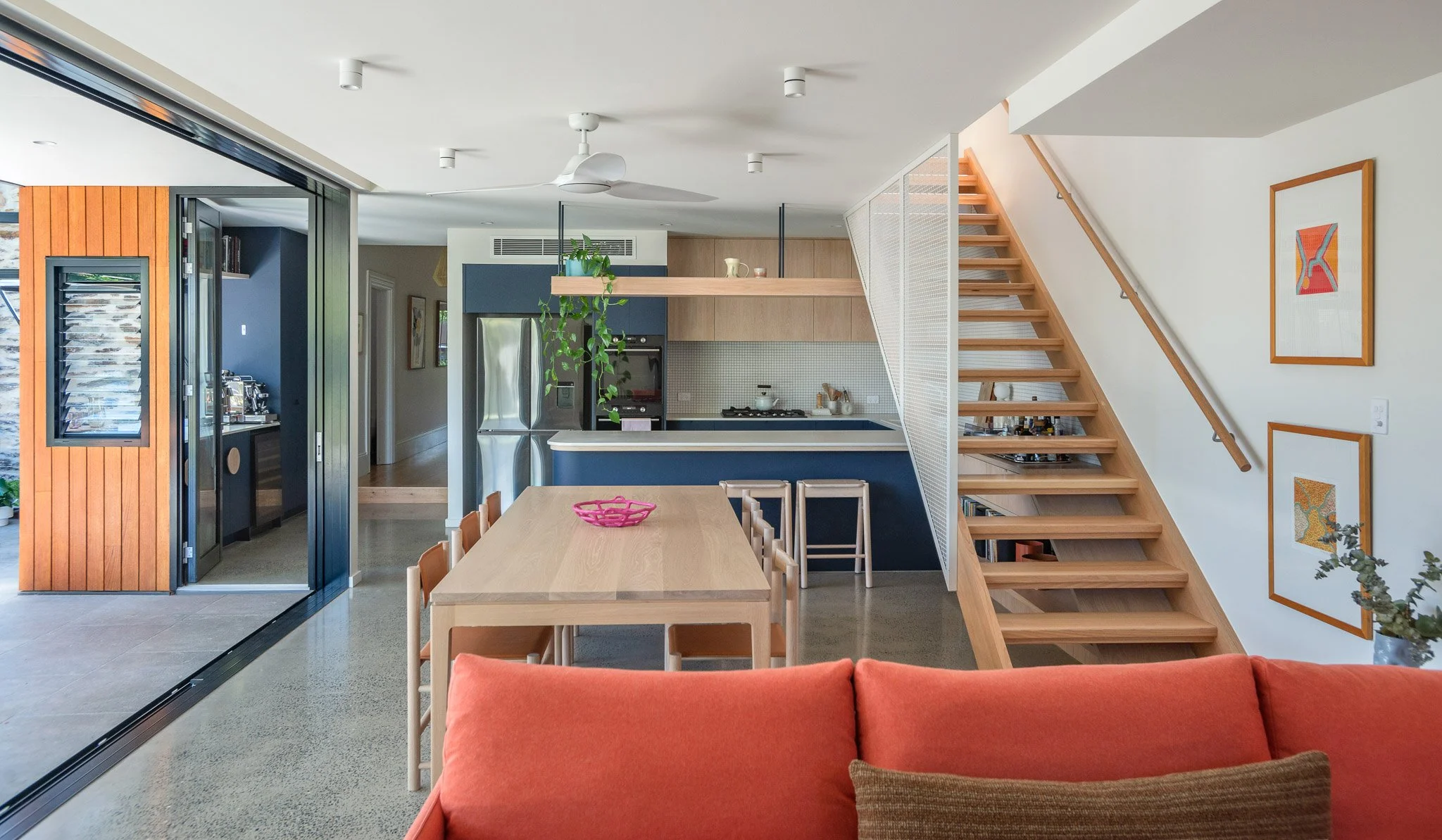 Interior of a modern home with a dining area and kitchen. A large wooden dining table with chairs is in the foreground, with a red sofa visible at the edge of the image. The kitchen features a blue base cabinet, a stainless steel refrigerator, and a wooden staircase leading to the upper floor on the right. A hanging shelf with green plants and decorations is above the kitchen. The walls are decorated with framed artwork, and the space has a minimalist, contemporary design.