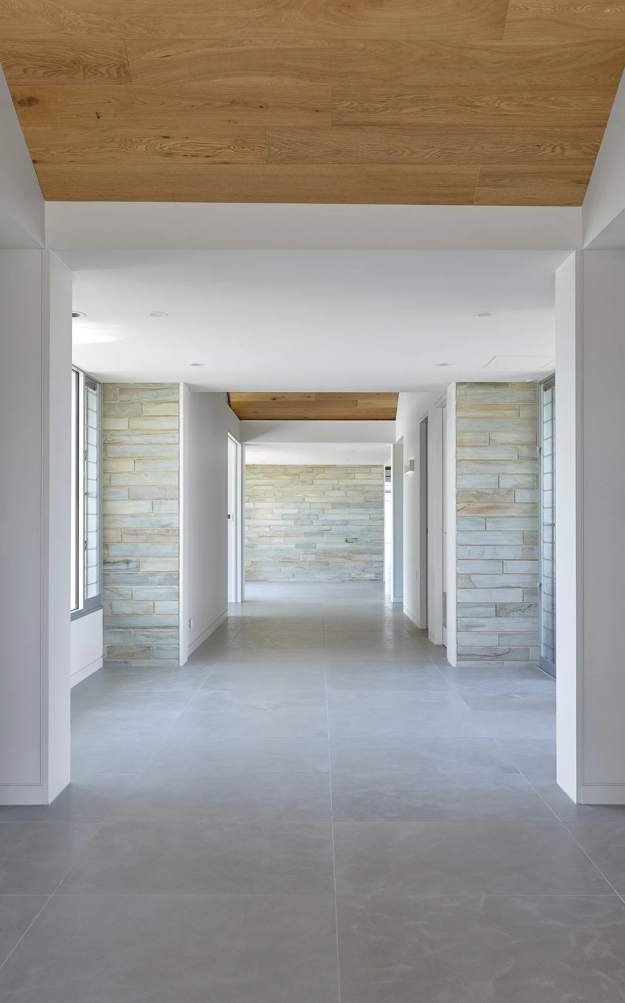 Modern hallway with light gray tiled floor, white walls, and stone accent walls, featuring a wood ceiling panel.