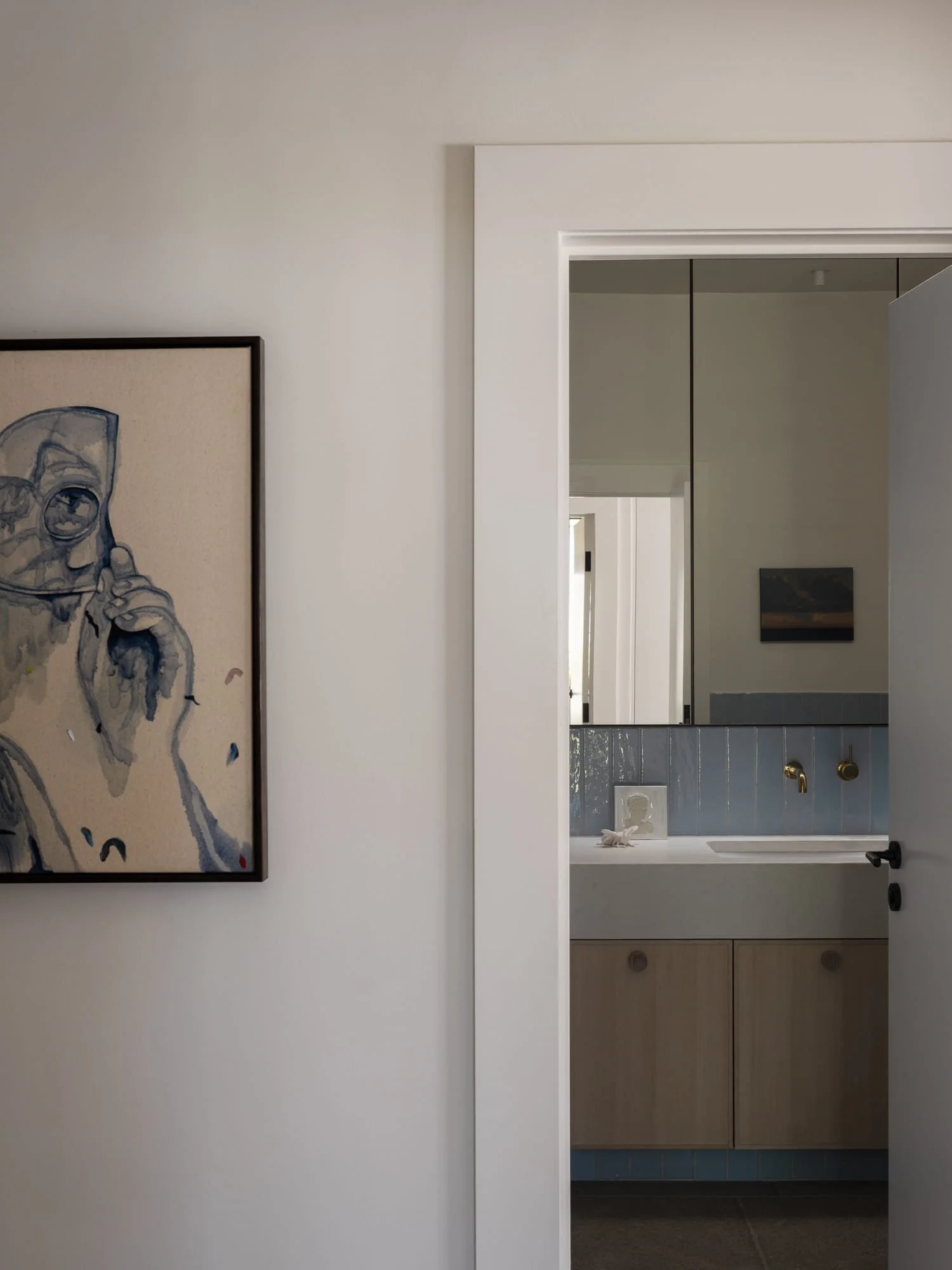 Interior view showing a hallway with artwork on the wall, leading into a bathroom with gray tiled walls and a wooden vanity.