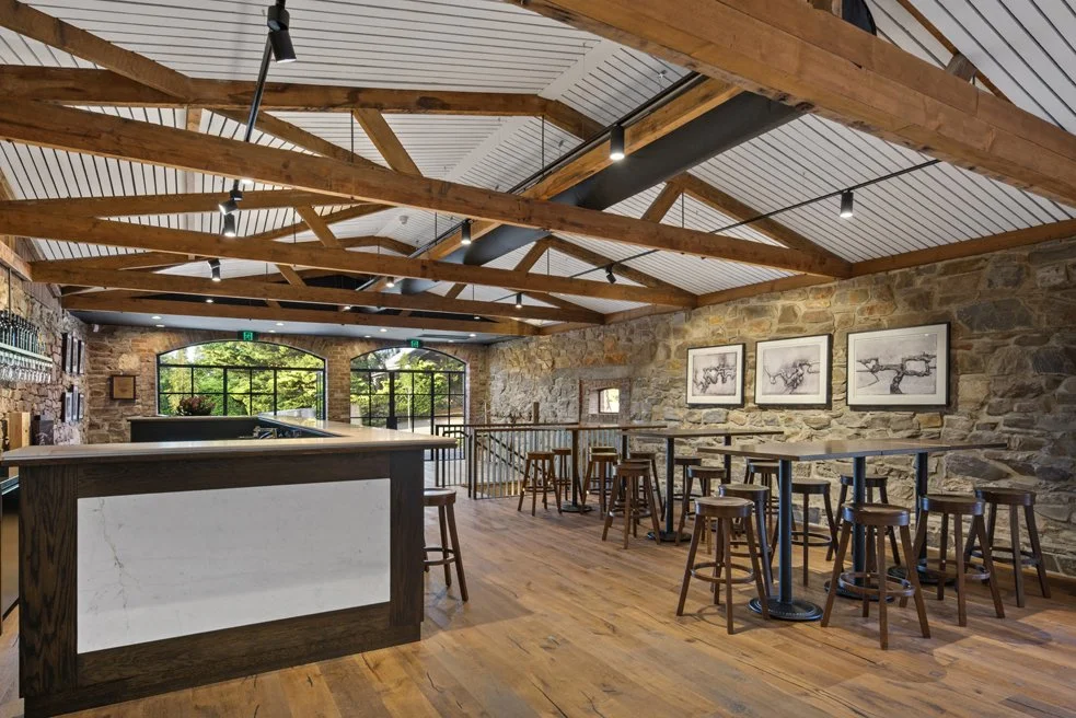 Interior of a rustic bar or cafe with wooden beams, stone walls, high tables with bar stools, and framed artwork hanging on the wall.