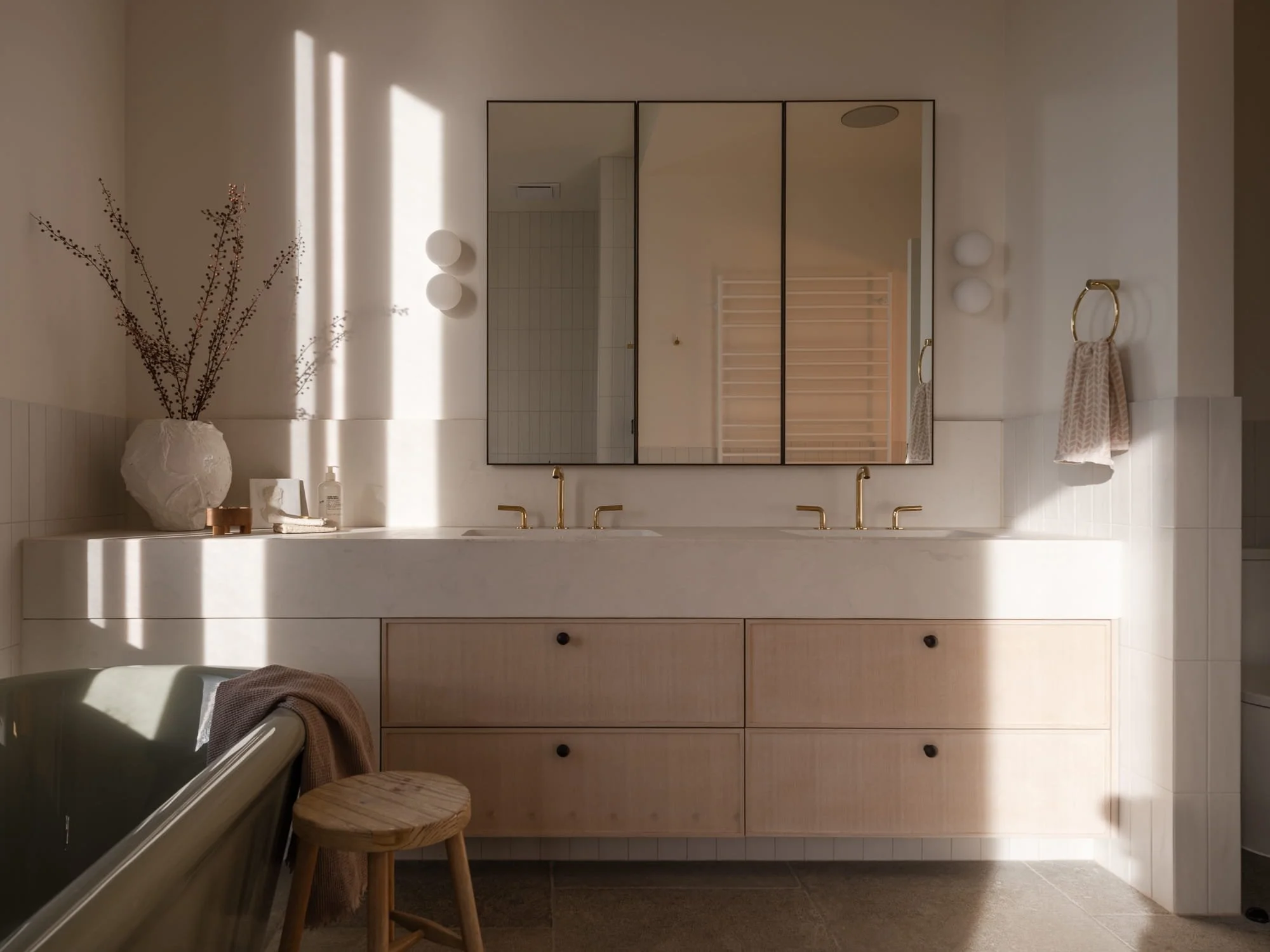 Minimalist bathroom with a large mirror above a white vanity, decorative branch in a white textured vase, and warm lighting fixtures.