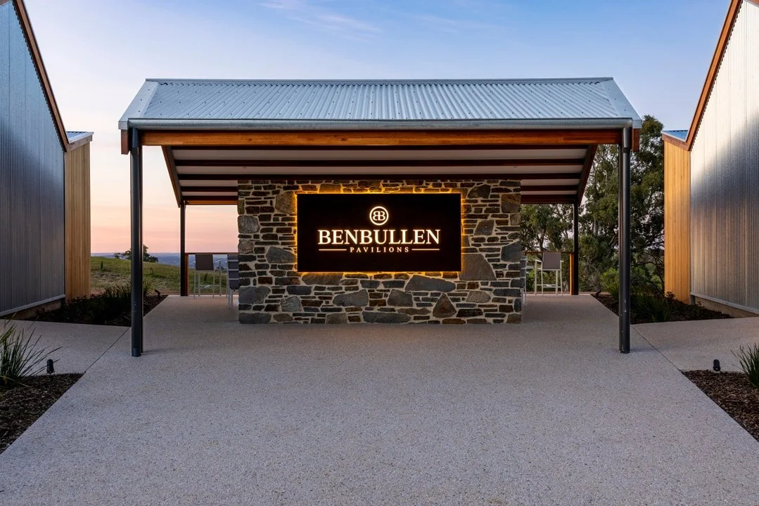 The entrance to Benbullen Pavilions features a stone wall with an illuminated sign displaying the name, under a metal roof supported by black poles, with a concrete pathway leading up to it, surrounded by landscaped plants and trees in the background.