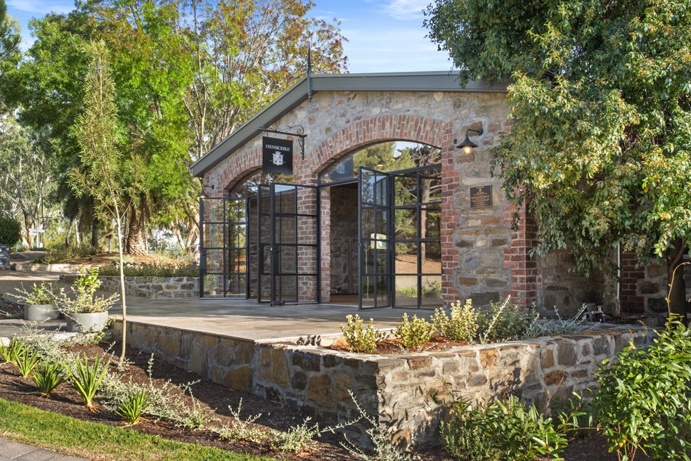 A small stone and brick building with large open glass doors, surrounded by trees and landscaped garden.