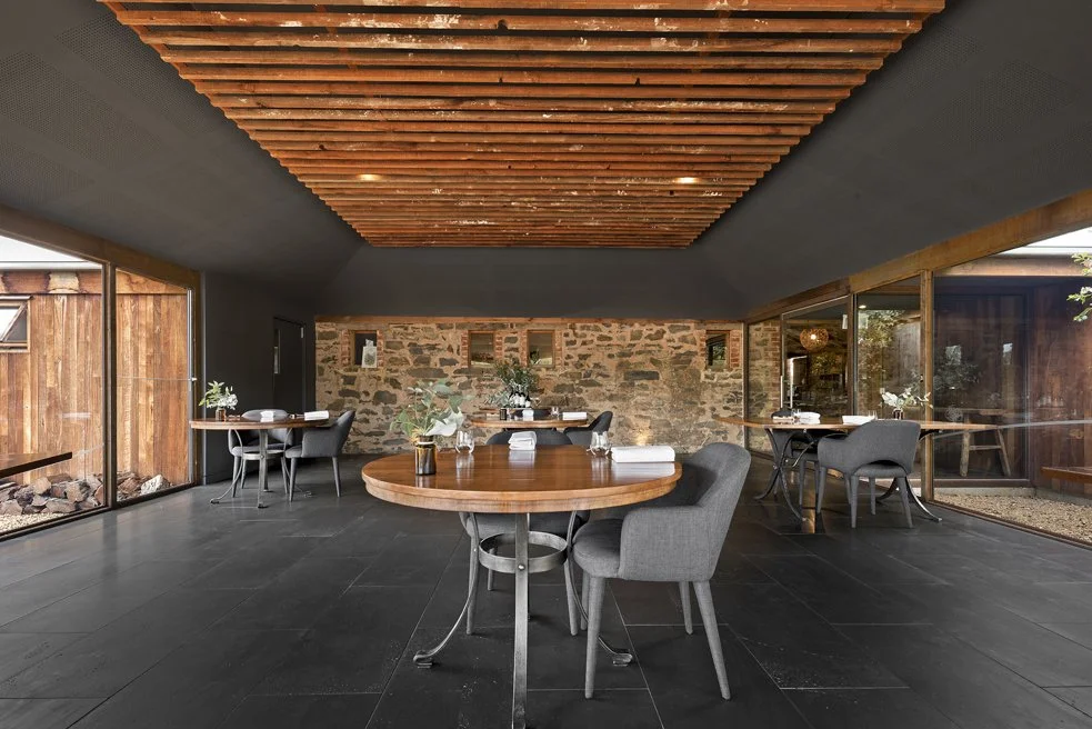 Modern dining area with dark tiled floor, wooden ceiling accents, stone wall, large windows, and simple furniture including round tables and gray chairs.