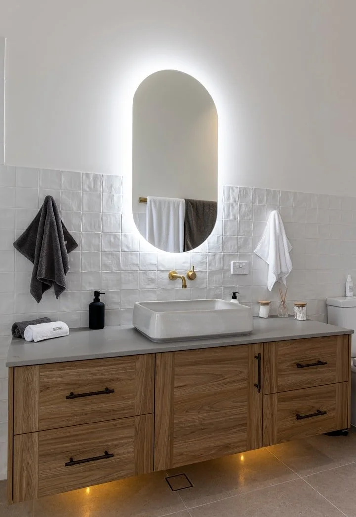 Modern bathroom with a vessel sink, gold faucet, and illuminated oval mirror, featuring wooden vanity, towels, soap dispenser, and toiletries.