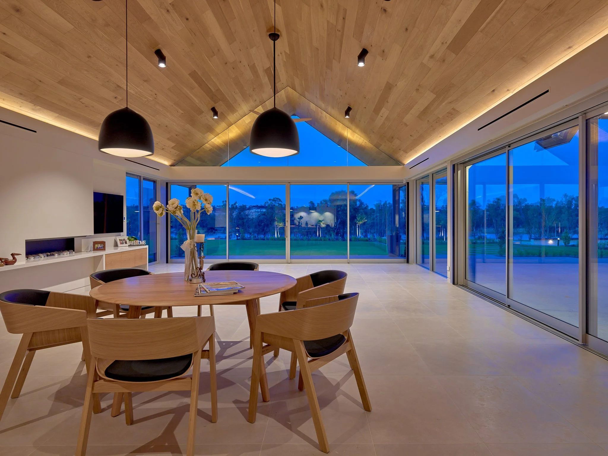 Modern living room with large windows, wooden ceiling, black pendant lights, and a round wooden dining table with black chairs.