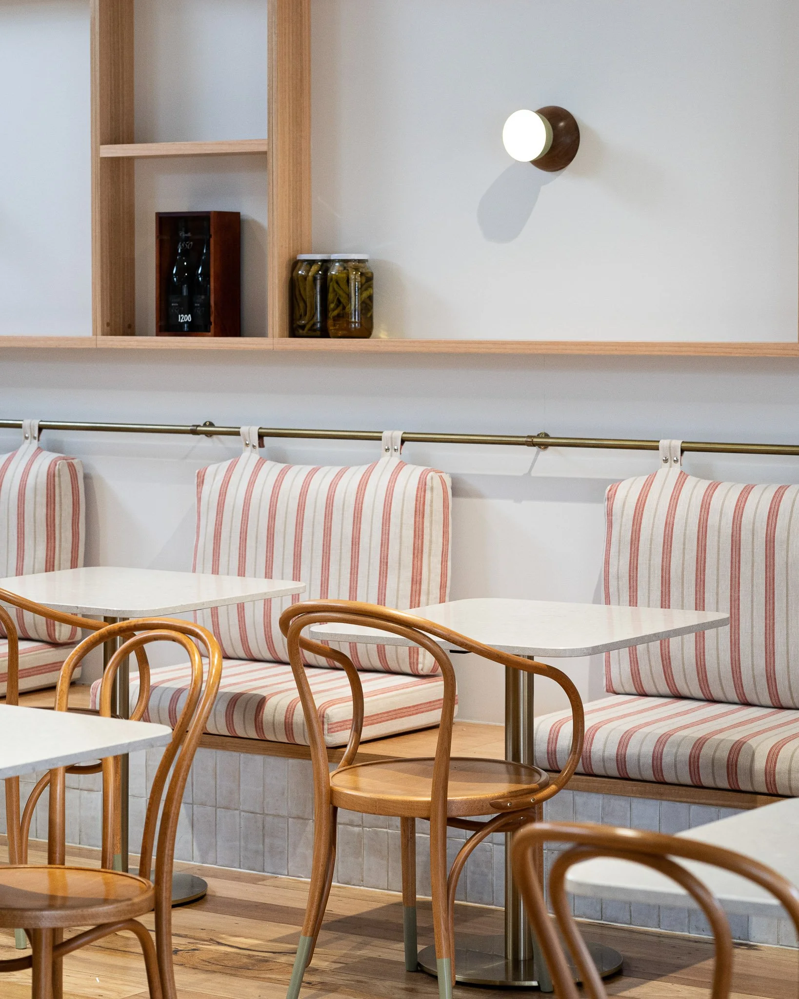 Interior of a cafe or restaurant with wooden chairs, round tables, striped cushioned bench seating, and a white brick wall with wooden shelf and jars.