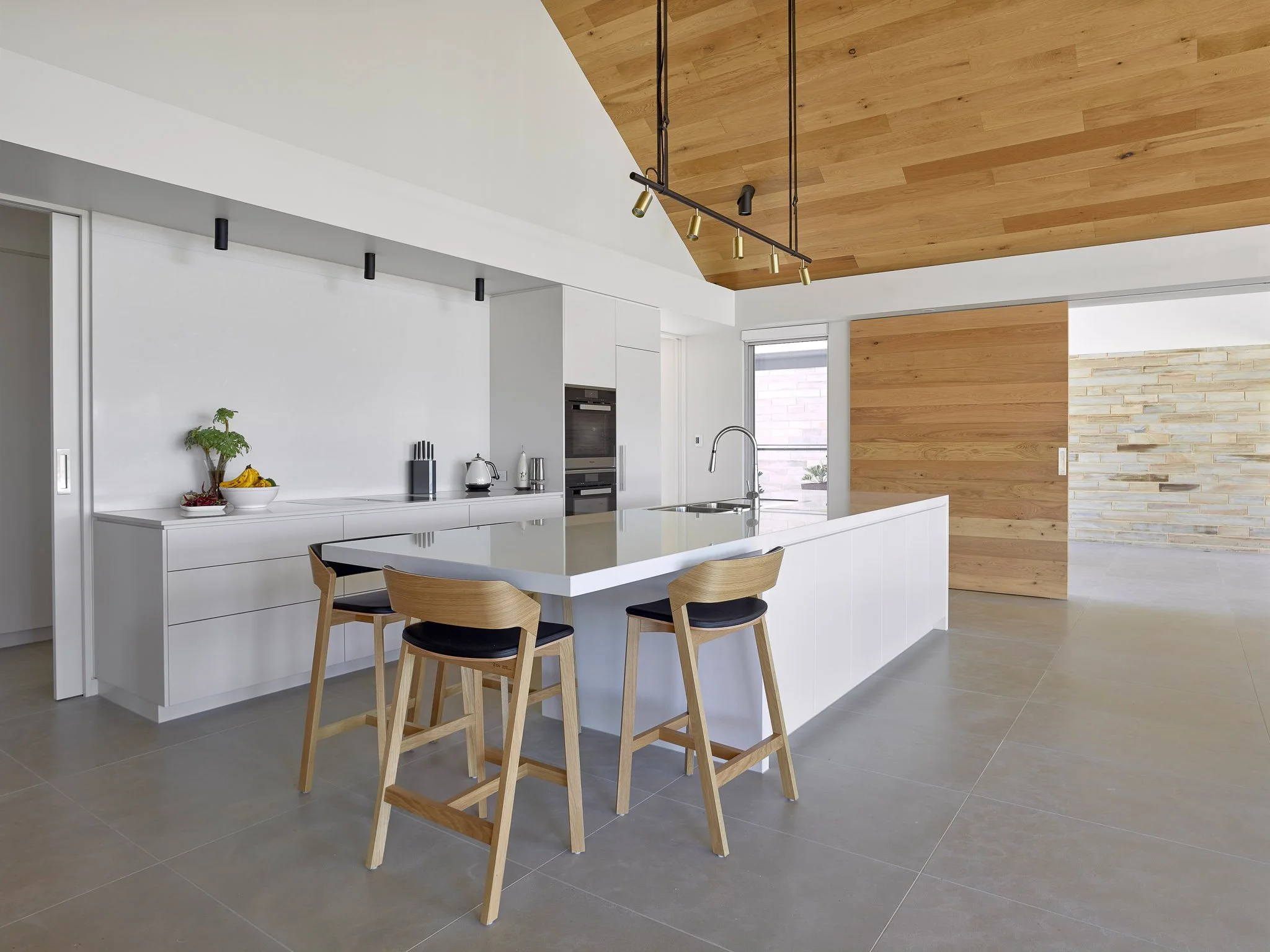 Modern kitchen with a white island, wooden chairs, and a wooden ceiling. There are black track lights and a bowl of bananas on the counter.
