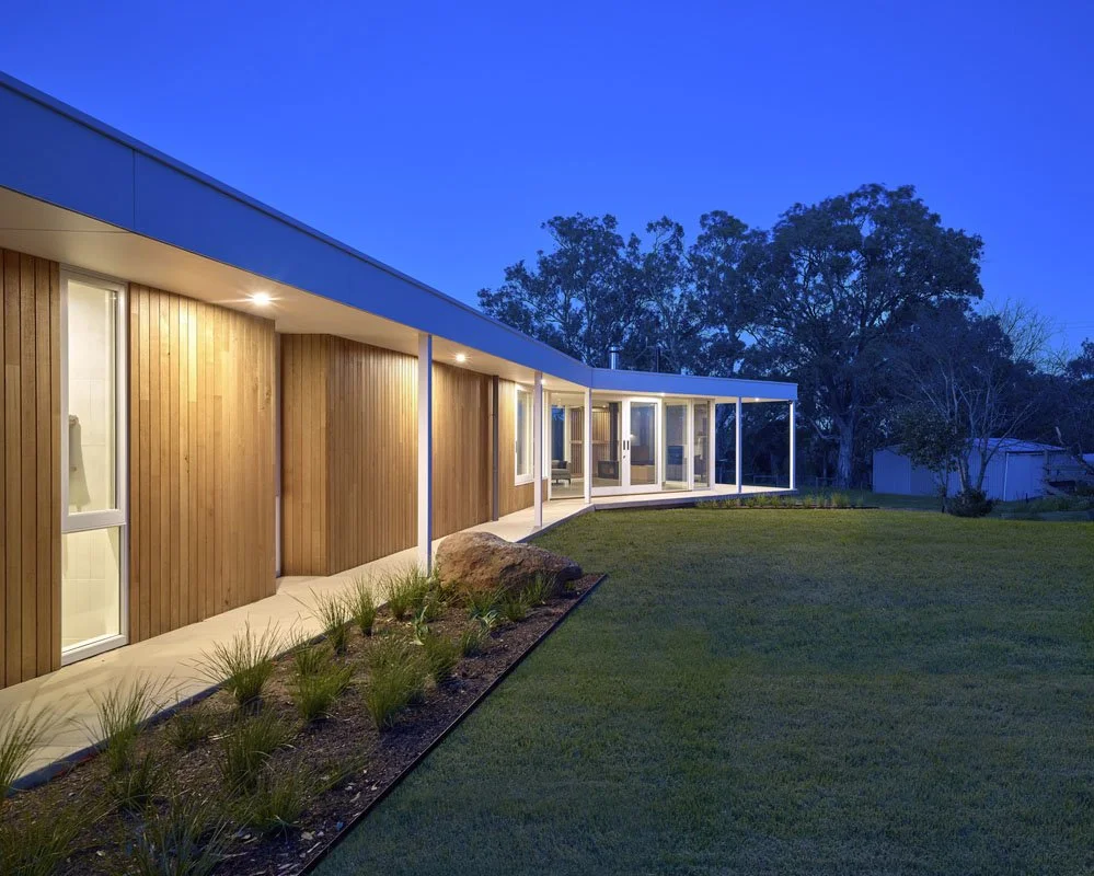 Modern house exterior at dusk with wooden panels, large glass windows, and a spacious grassy yard.