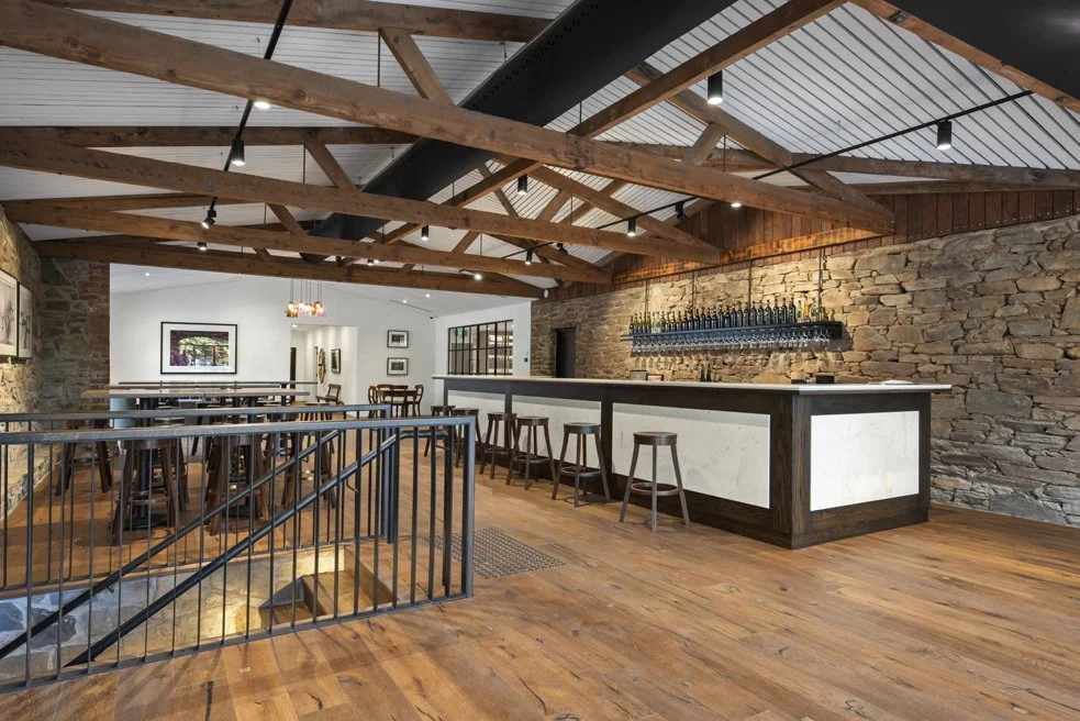 Interior of a rustic-style bar and restaurant with wooden beams, a stone wall, and a white bar counter with stools.