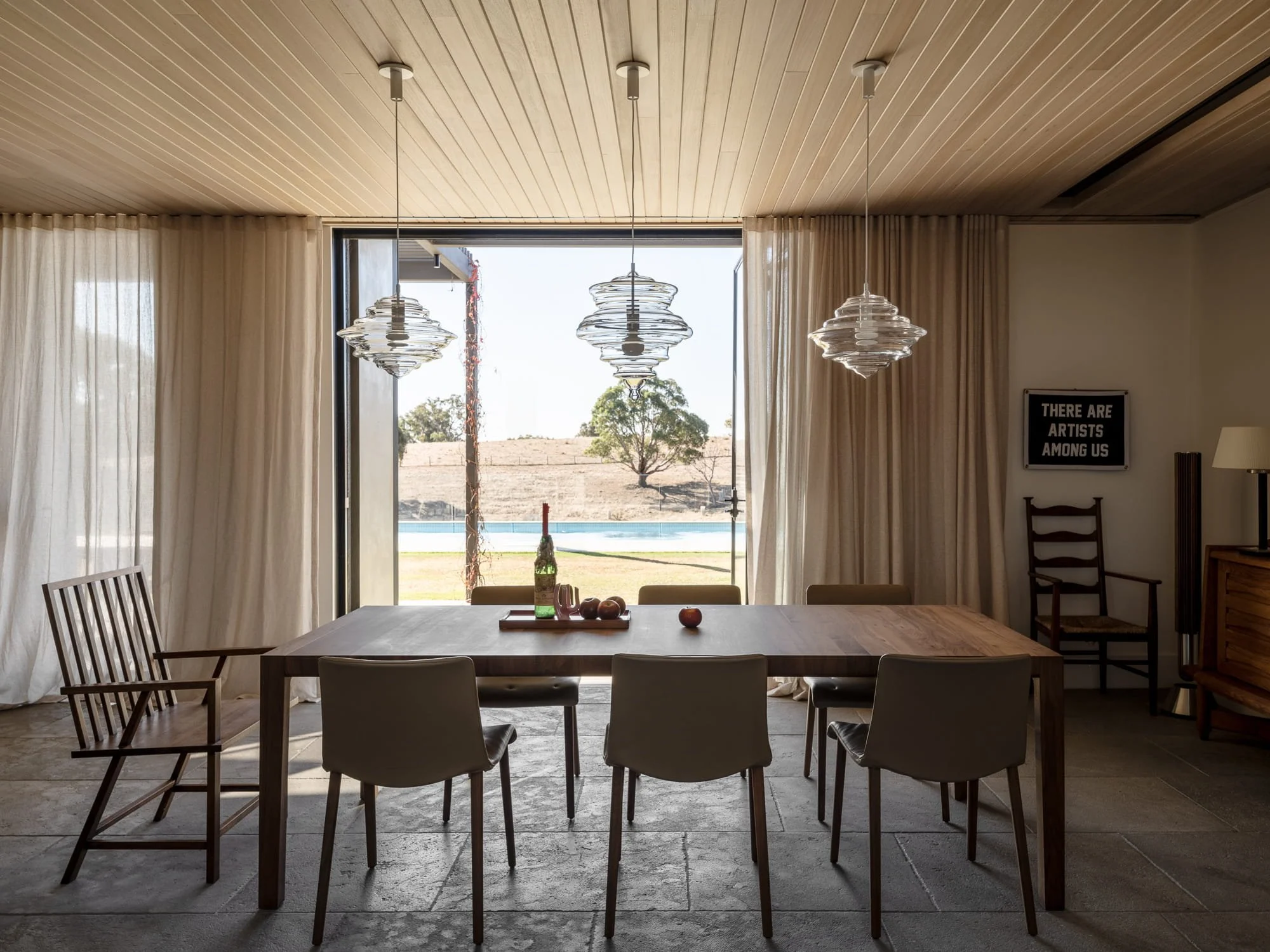Dining room with wooden ceiling, three pendant lights, large open sliding glass door, white curtains, and a view of a tree and fields outside.